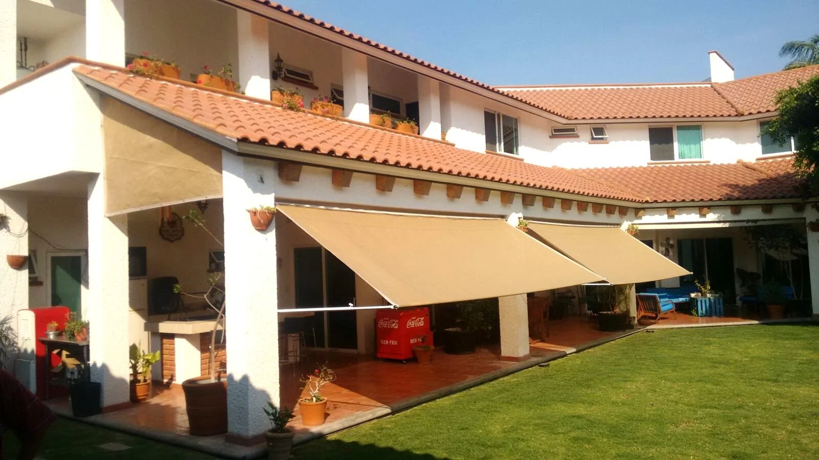 Casa de varias plantas con terraza, toldos beige, paredes blancas, tejas rojas, jardín con césped y plantas en macetas y varias mallas personalizadas