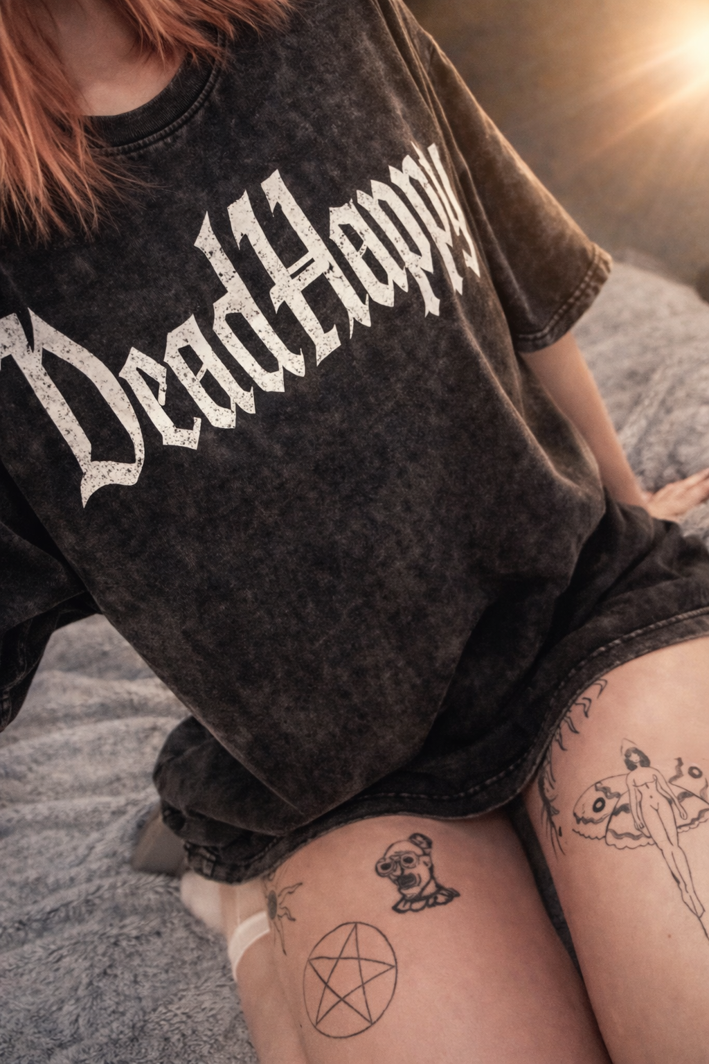 Close-up Of A Person Wearing A Black T-shirt With The Word Dead Happy Printed In White, View Of Their Bare Legs With Tattoos Including A Female Figure, A Pentagram, And A Butterfly, Sitting On A Grey Plush Surface.