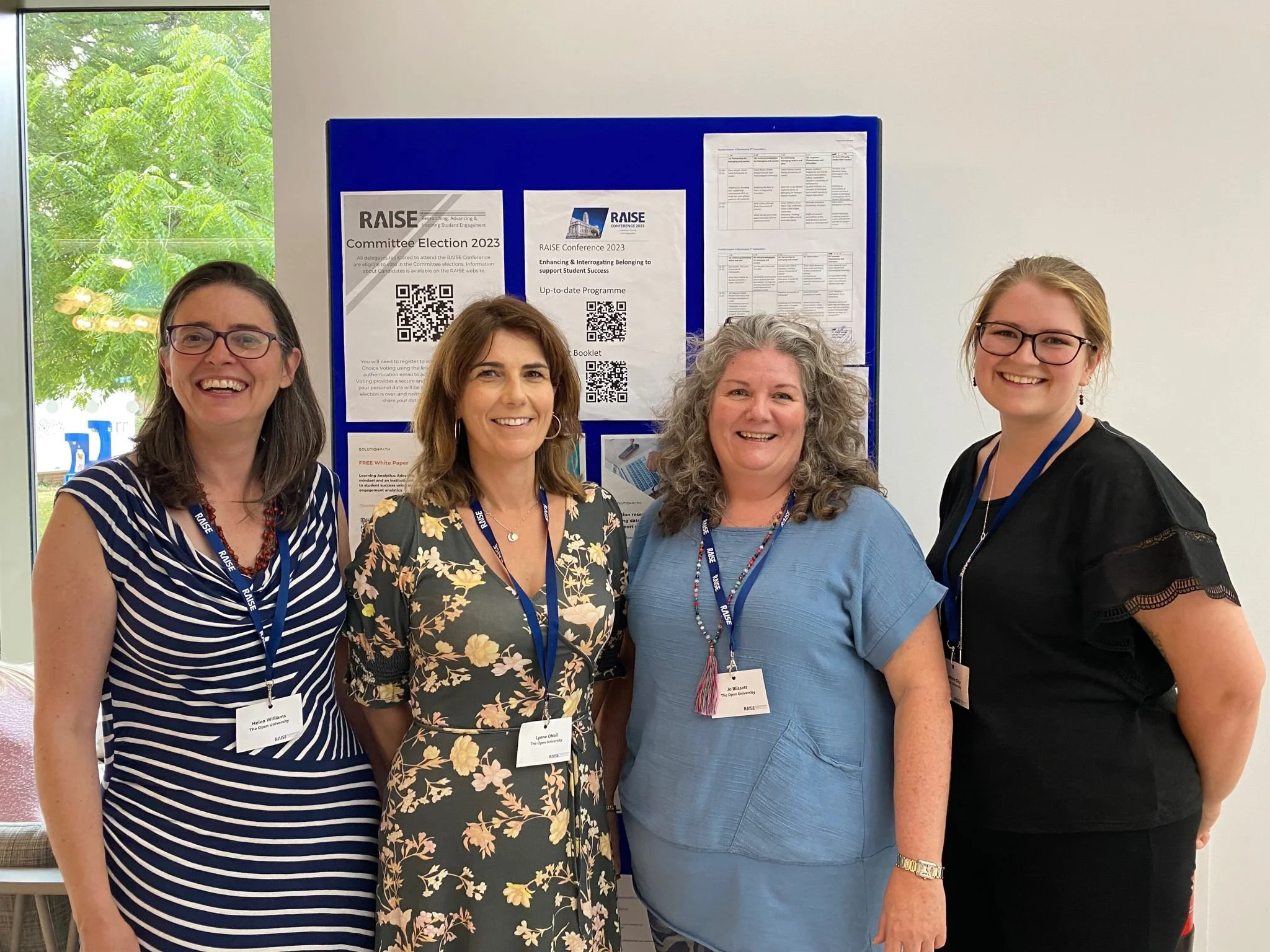 Four women standing in front of a blue bulletin board with posters and flyers, smiling at the camera at a conference event.