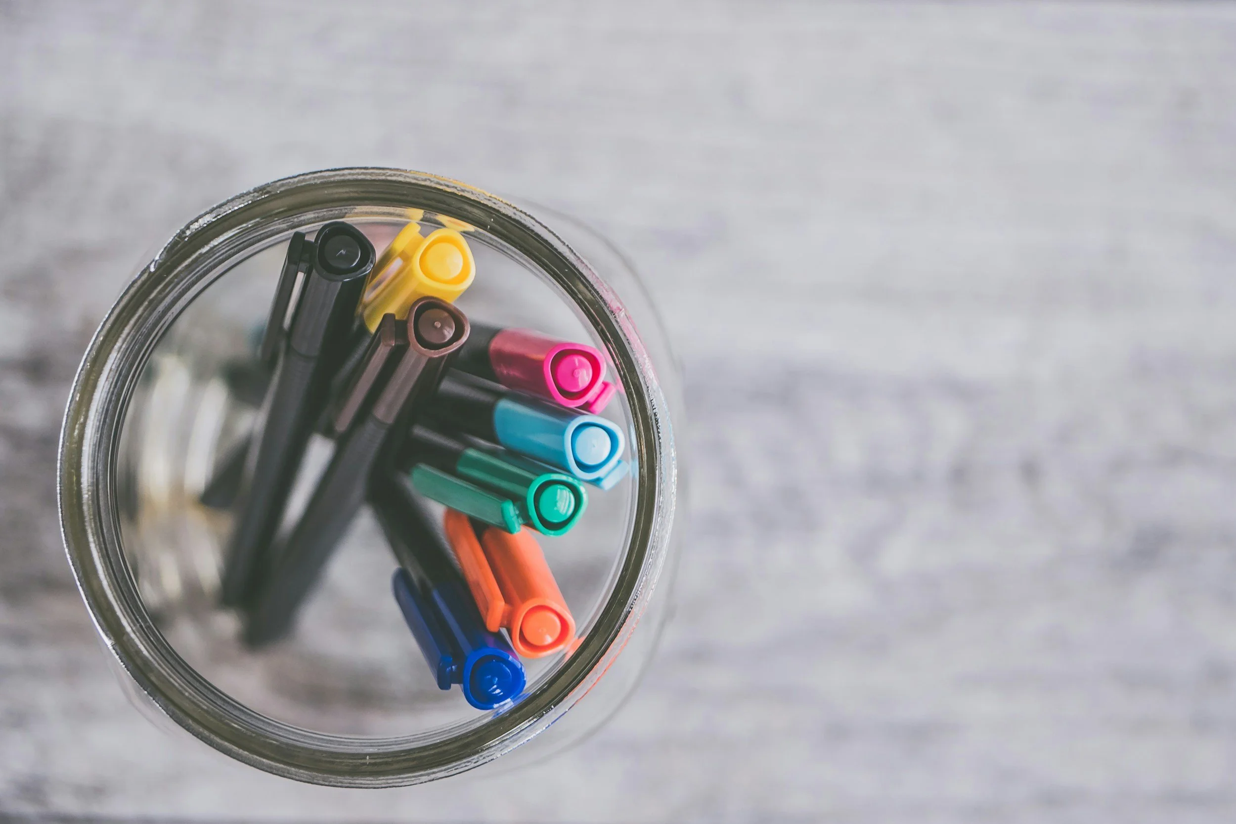 A top-down view of a clear glass jar filled with multicolored felt-tip pens arranged in a circle on a light gray wooden surface.