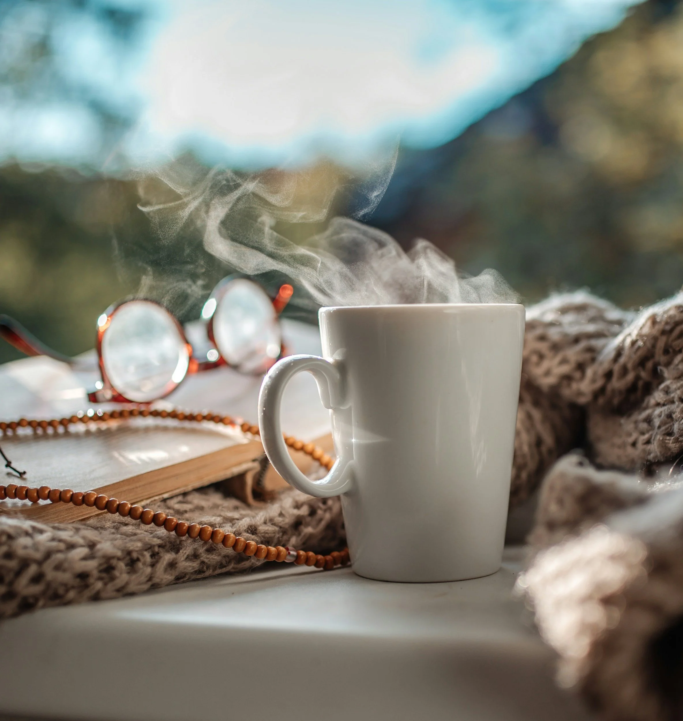 A white mug with steam rising from hot beverage, sunglasses, an open book, and a beaded necklace on a soft blanket, with blurred greenery outside in the background.