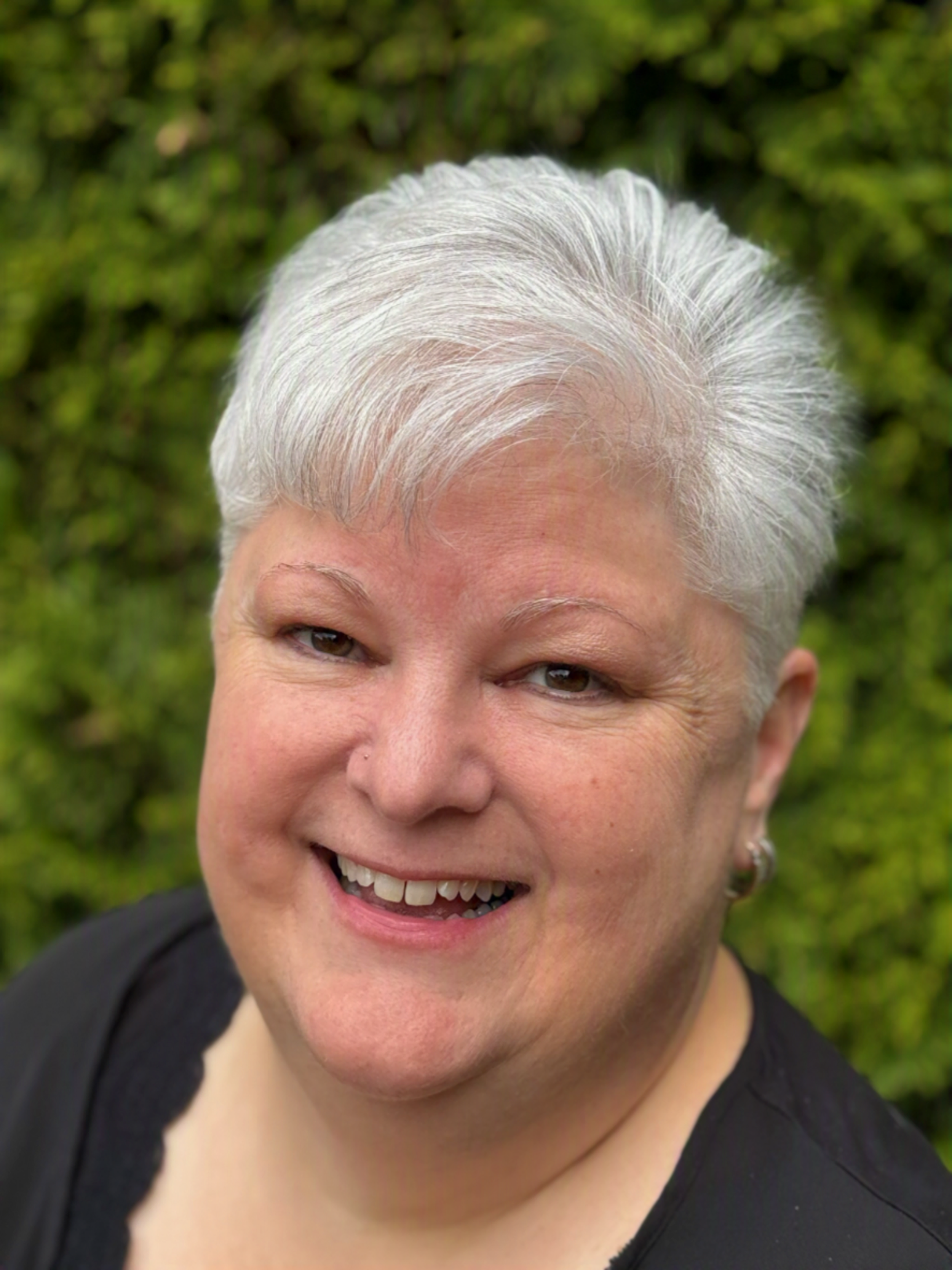 Close-up of Lauren K. Harris with short, white hair, smiling, and wearing a black top. Background is green foliage.