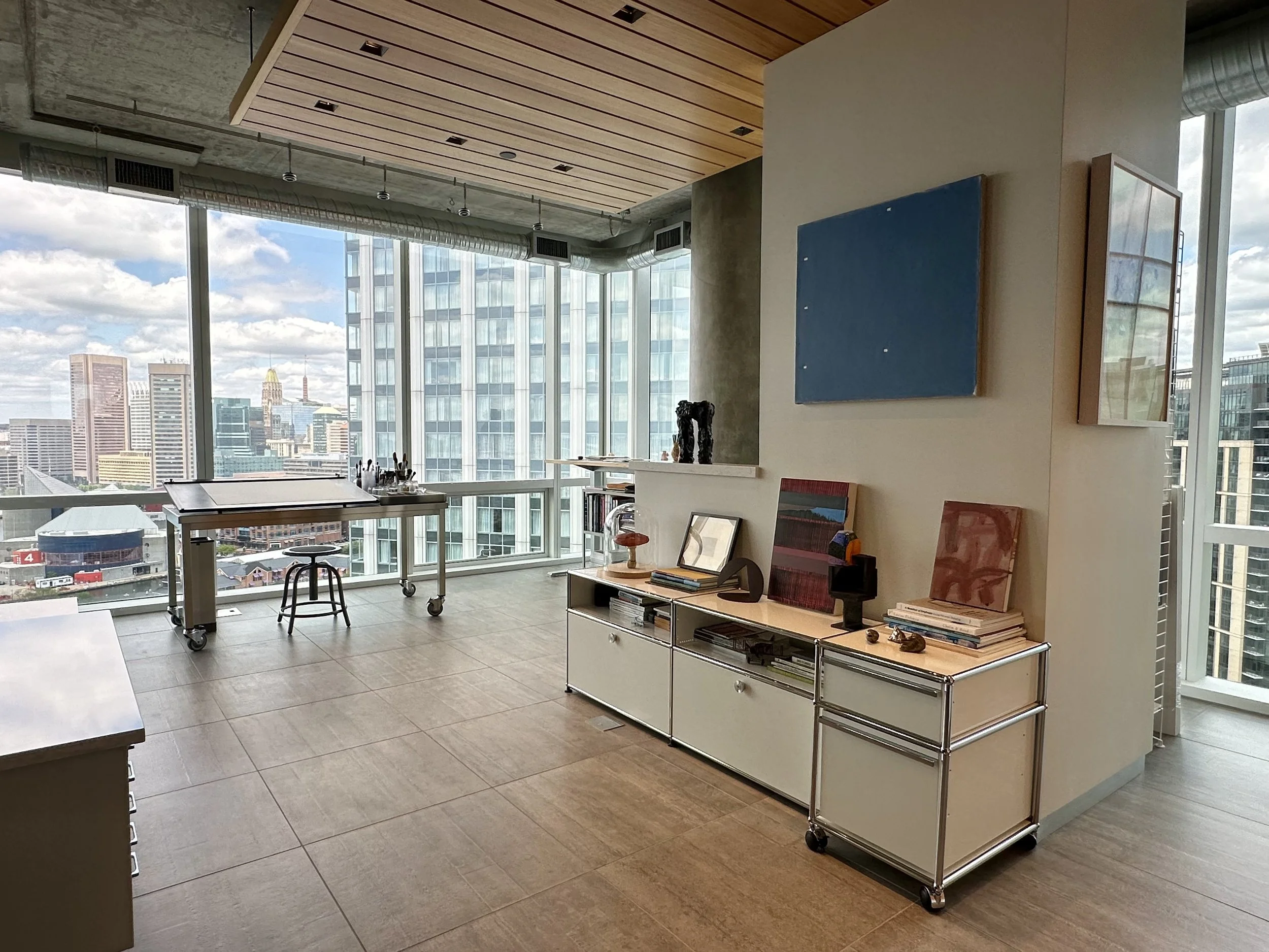 Modern art studio with large floor-to-ceiling windows, city skyline view, wooden ceiling, and art supplies on a desk.