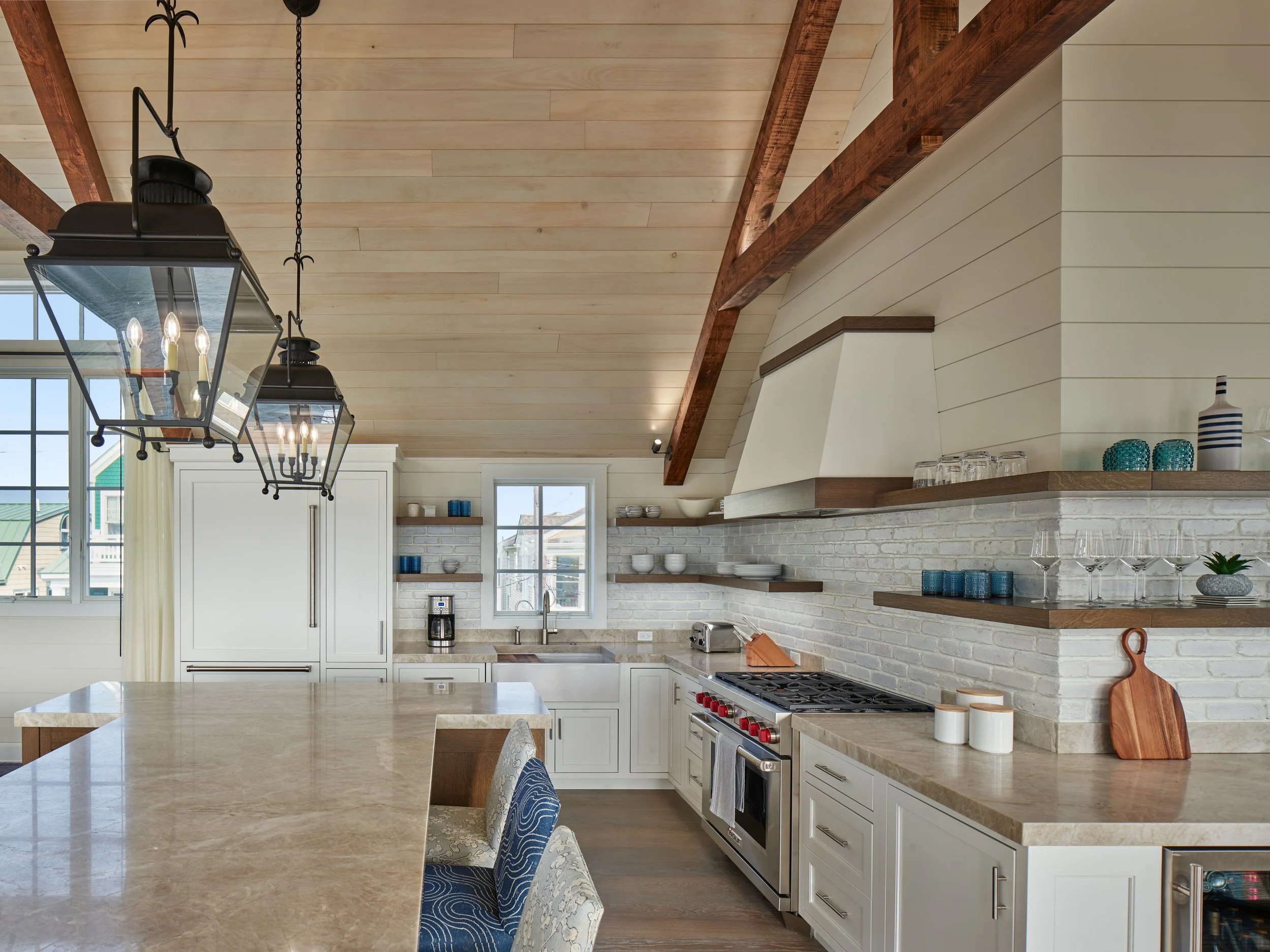 Modern kitchen with white cabinets, beige countertops, brick backsplash, and open wooden shelves. A window and a large kitchen island with seating are visible. Pendant lights hang above the island.