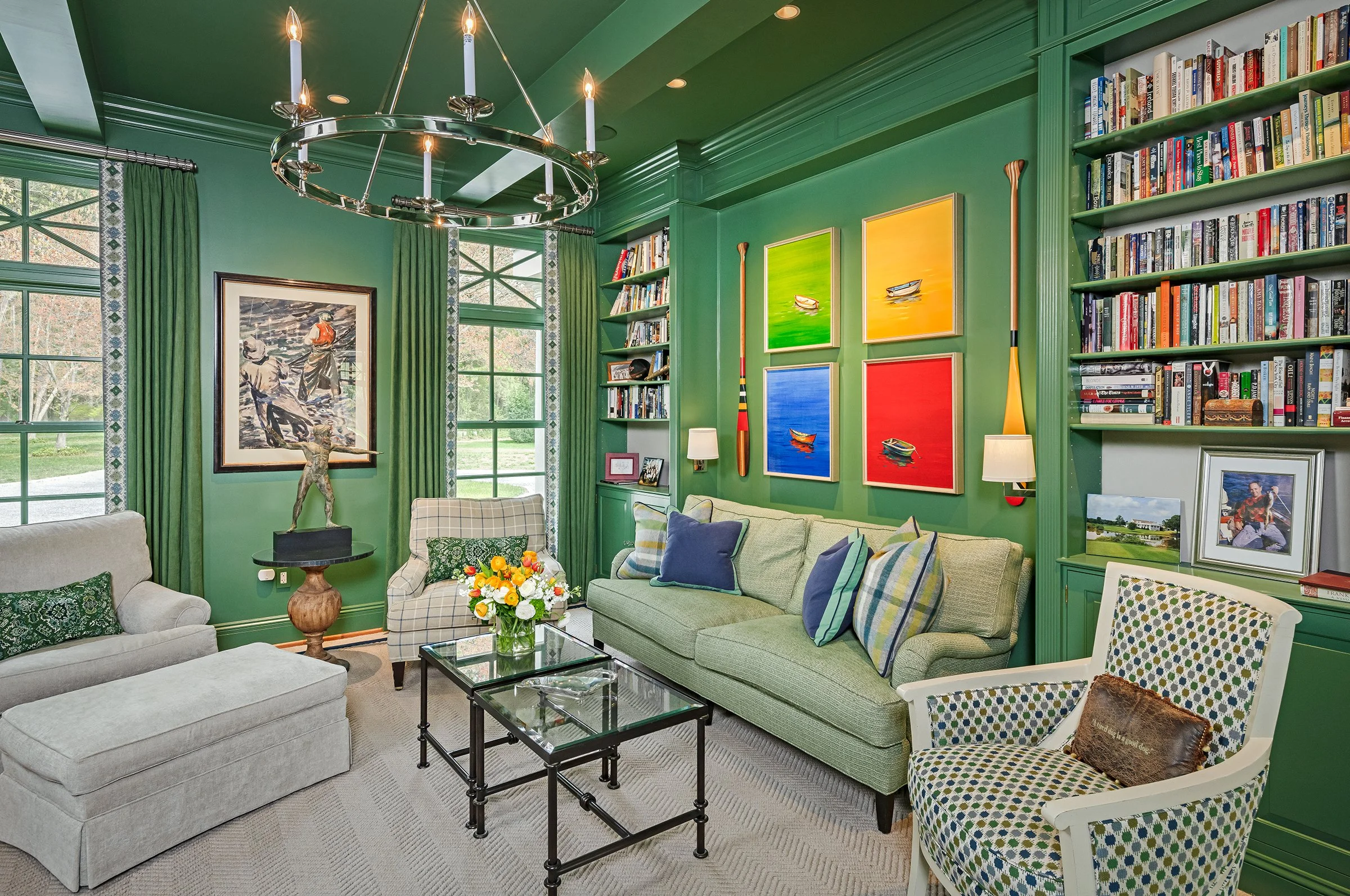 A living room with green walls, large windows, a chandelier, a sofa with blue and plaid pillows, a patterned armchair, a coffee table with a flower arrangement, bookshelves filled with books, four colorful paintings of boats, and various framed pictu