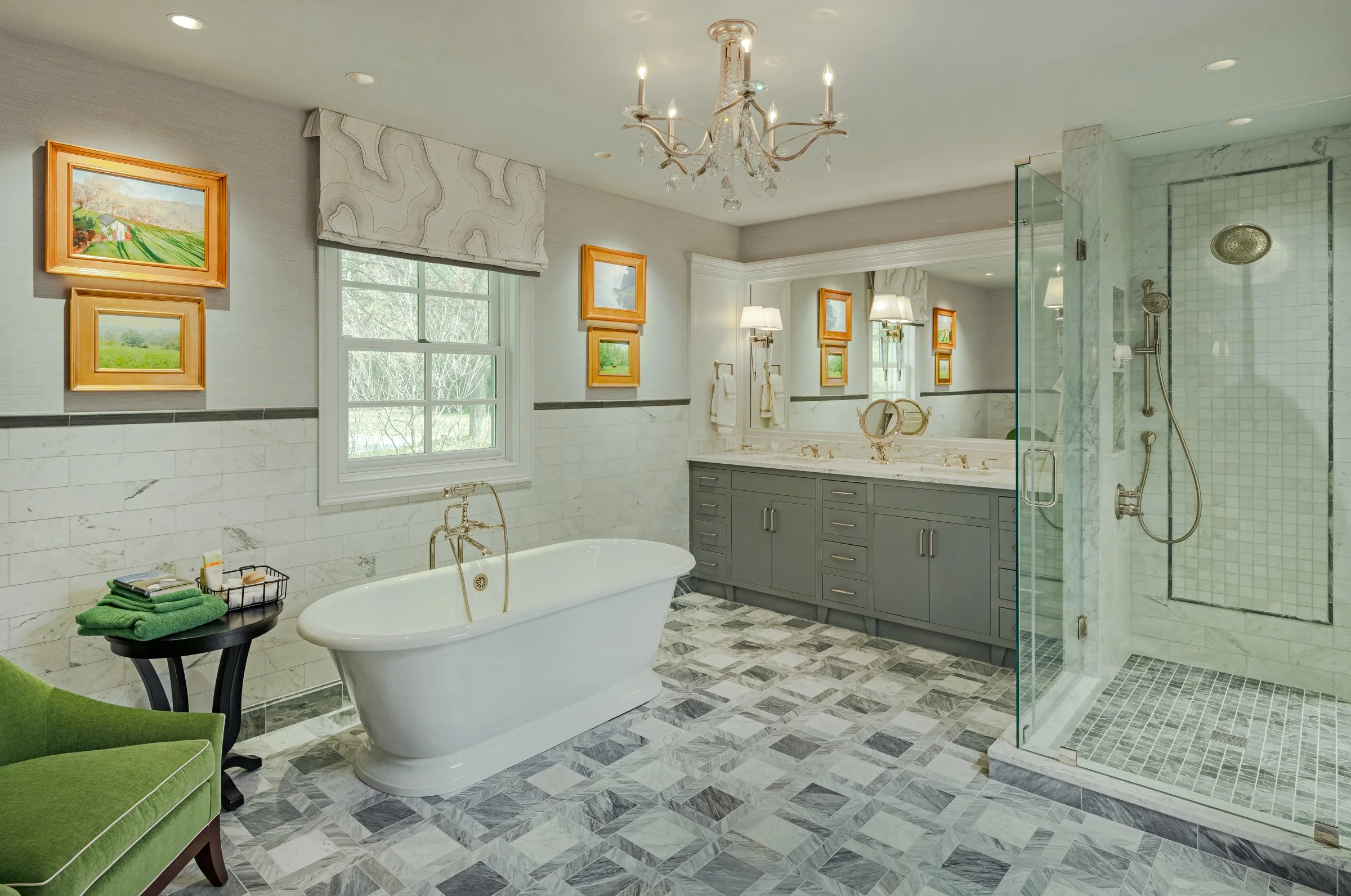 Luxury bathroom with a white clawfoot bathtub, gray vanity with marble countertop and dual sinks, glass-enclosed shower with marble walls, framed landscape paintings, green chair, window with a patterned white valance, chandelier, and patterned tile 