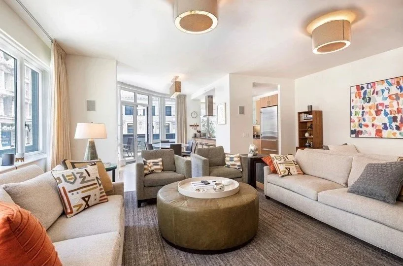 Brightmodern living room with multiple sofas, a round ottoman, colorful throw pillows, a large window with beige curtains, and artwork on the wall.