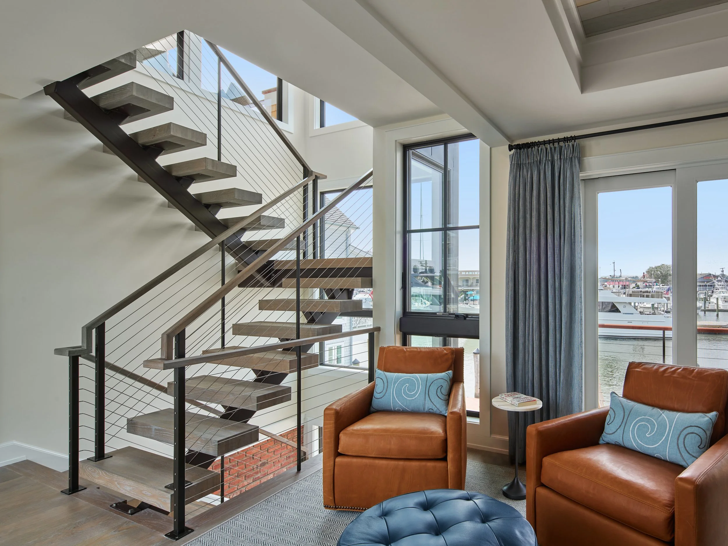 Living room with orange leather armchairs, a small side table with a magazine, large windows overlooking a marina with boats, a staircase with wooden steps and metal railing, and blue curtains.