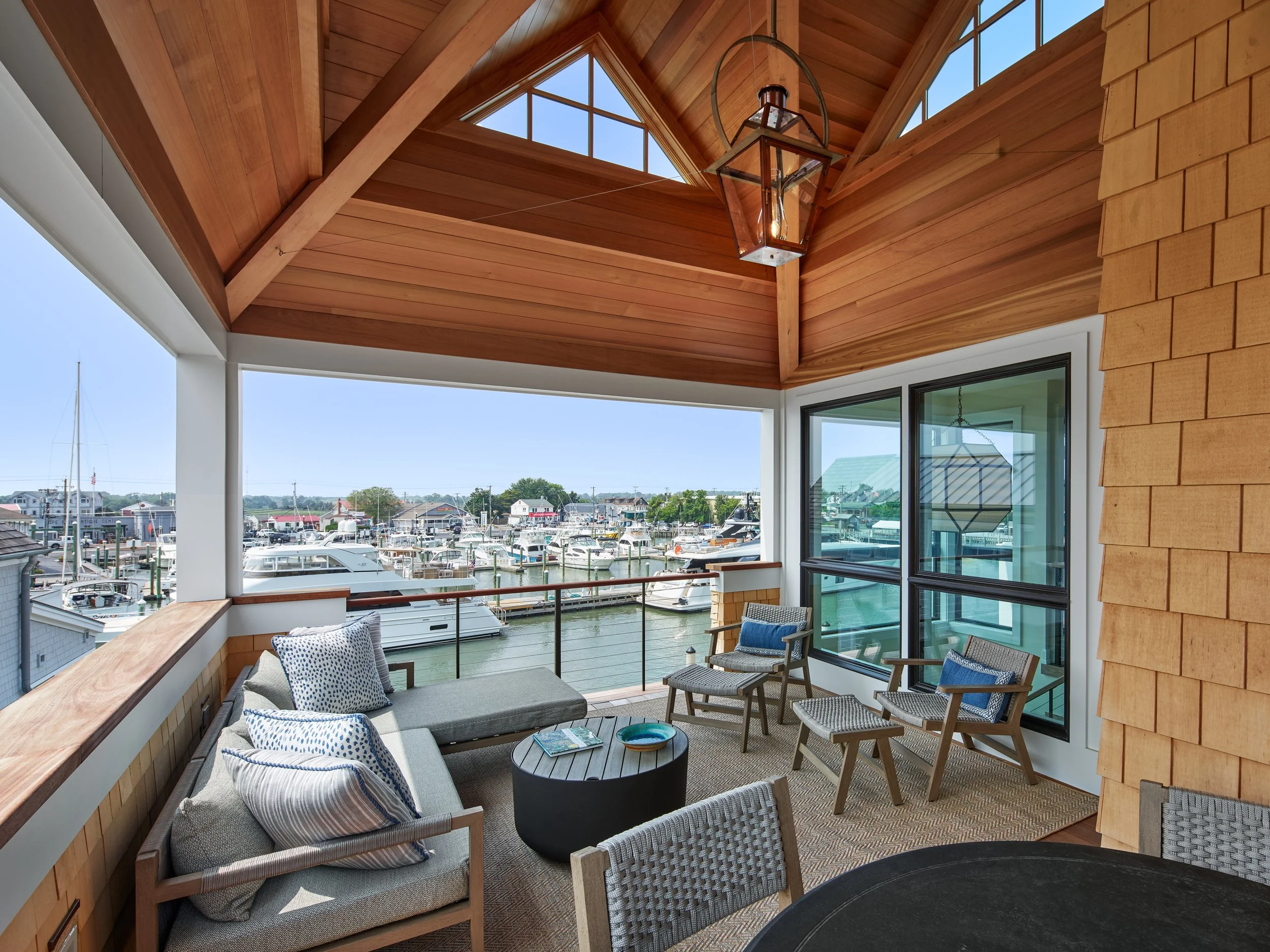 A covered porch overlooking a marina filled with boats, featuring wooden ceiling, glass windows, and outdoor seating with chairs and a round table.