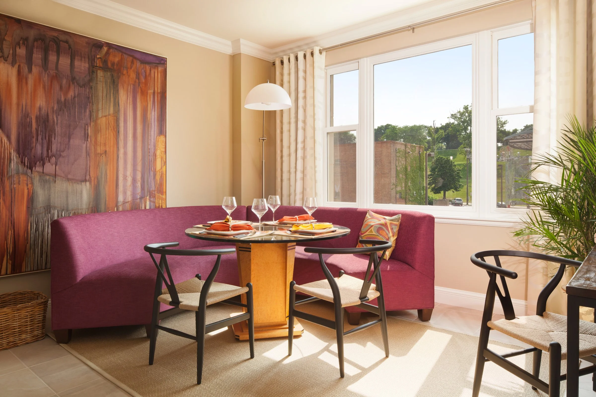 Dining area with a round table set with four wine glasses and colorful napkins, surrounded by a pink curved banquette and black chairs, with large windows letting in natural light and a view of green trees and buildings outside.