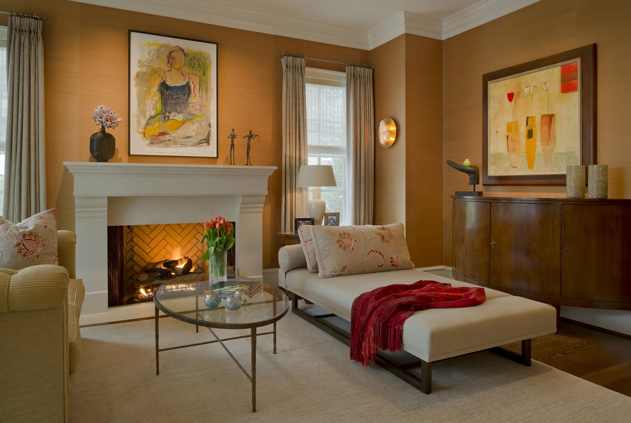 Living room with a fireplace, beige sofa and chaise lounge, a glass coffee table with tulips, framed abstract artwork, and various decorative items, warm lighting.
