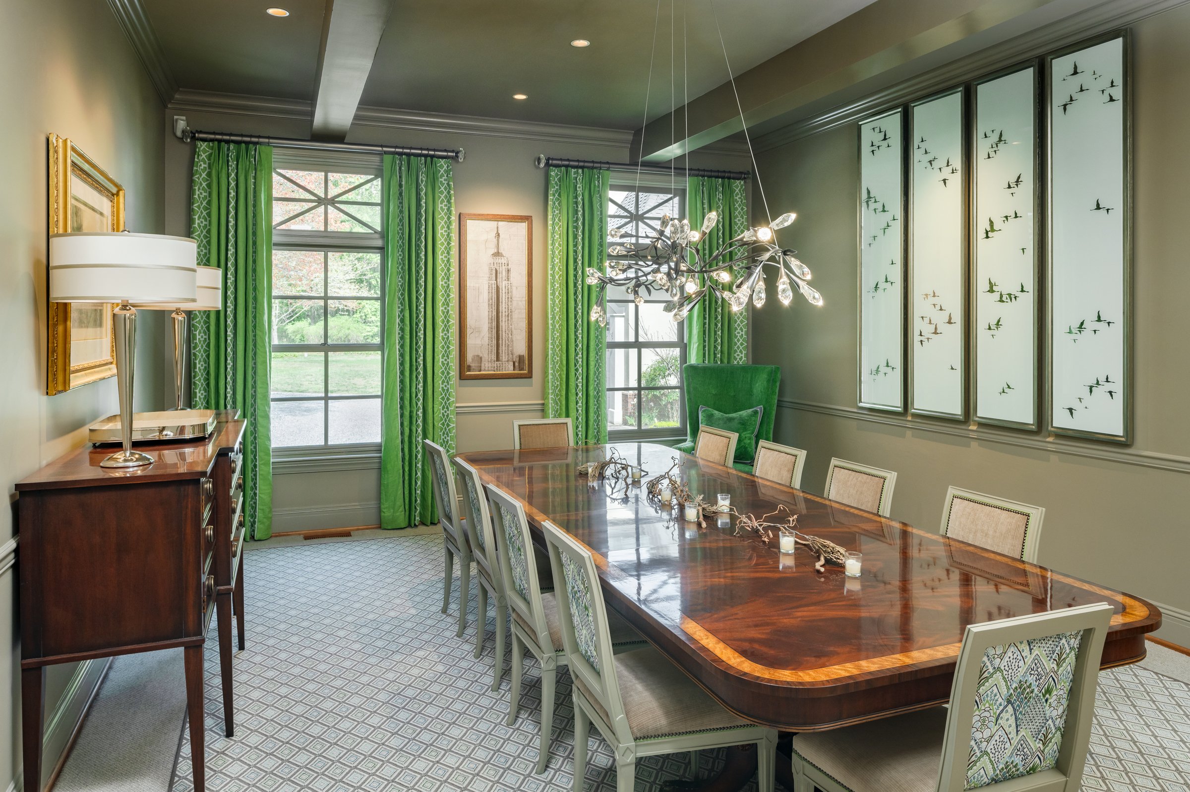 A bright dining room with a large wooden table surrounded by eight upholstered chairs, green curtains on two windows, a sideboard with a lamp, framed artwork, and a modern chandelier, with a 3-panel art piece on the wall depicting flying birds.