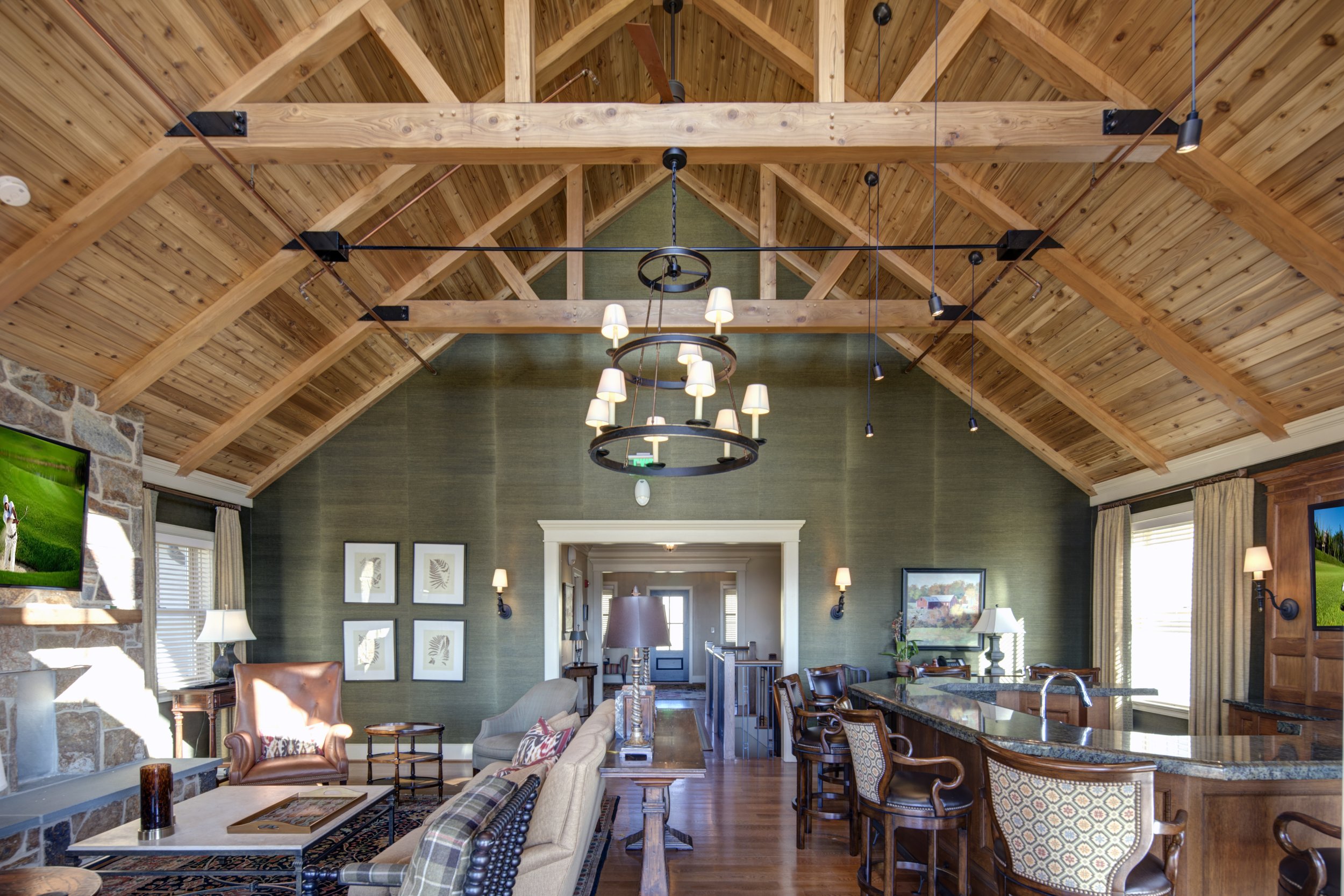 Living room with vaulted wooden ceiling, green accent wall, and rustic decor. Features a chandelier, wall sconces, a stone fireplace, flat-screen TVs, and a kitchen island with bar stools.