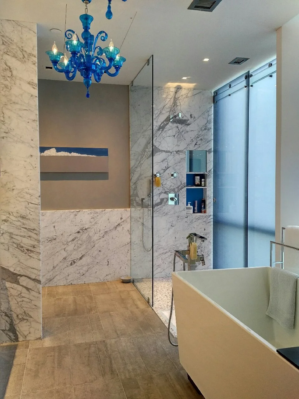 Modern bathroom with a blue chandelier, marble walls, a walk-in shower with shelves, a bathtub with a towel, and a large frosted glass door.