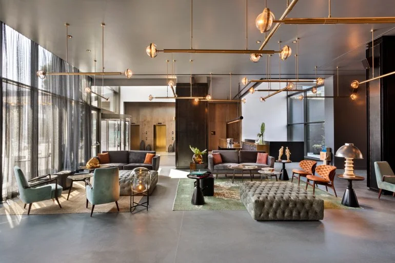 Modern hotel lobby with contemporary furniture, gold lighting fixtures, large floor-to-ceiling windows with curtains, and sleek decor.