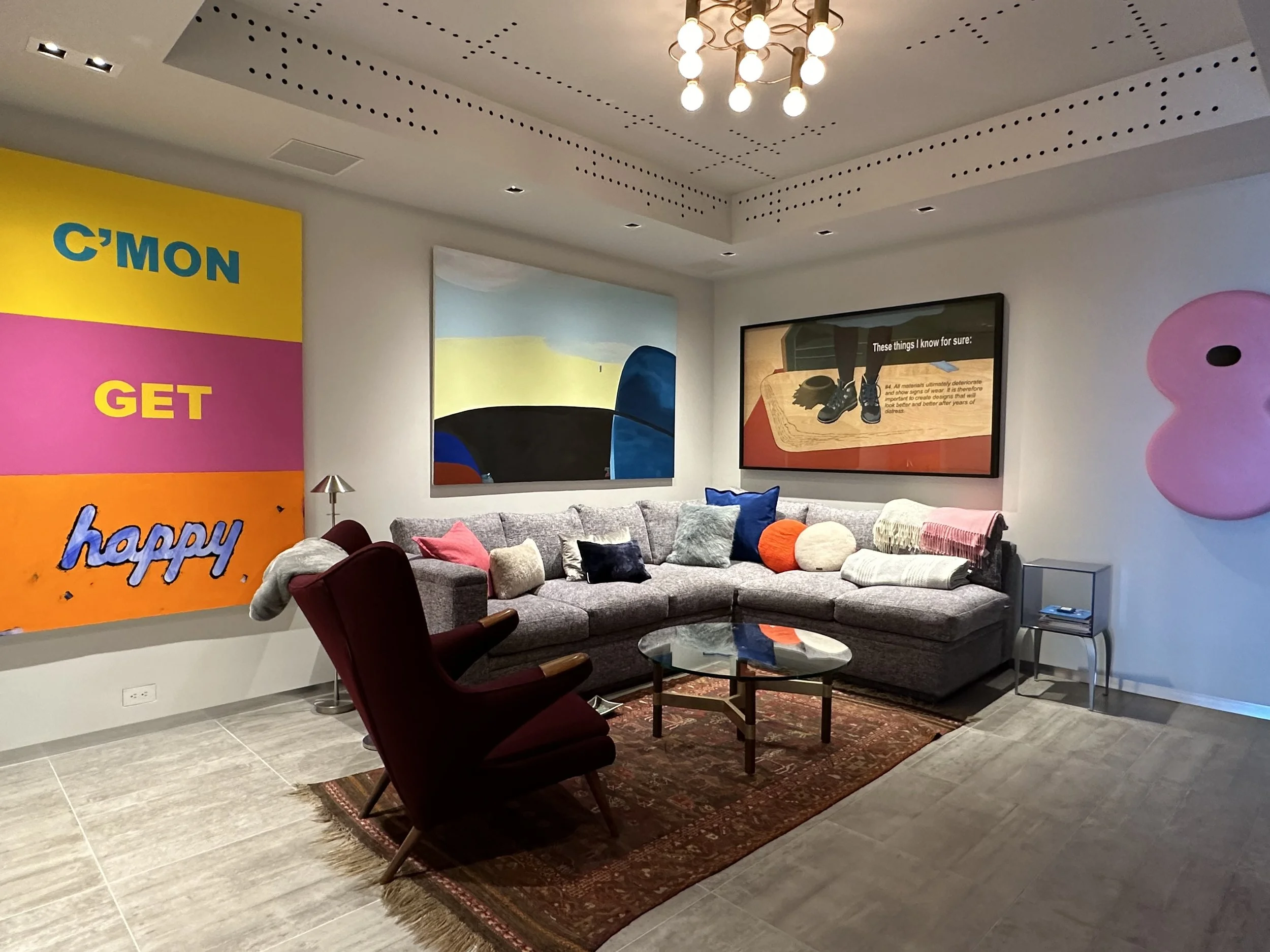 A contemporary living room with a large gray sectional sofa decorated with colorful pillows, a glass coffee table on a patterned rug, red armchair, artwork on the walls, and a modern ceiling light fixture.