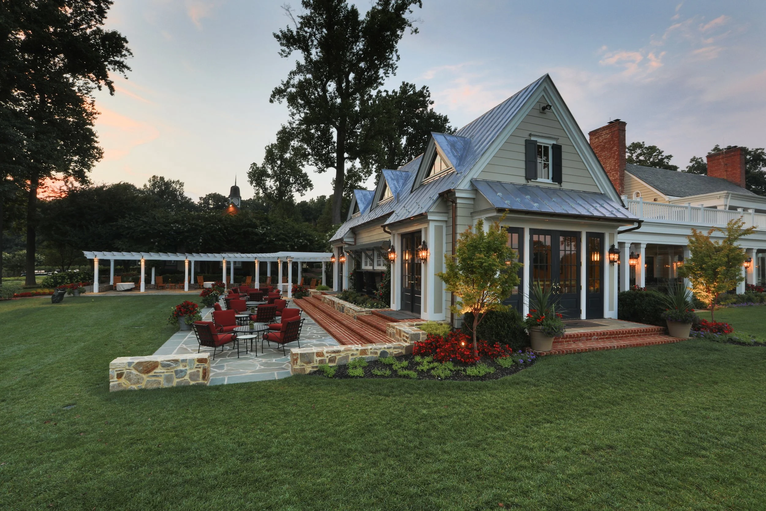A large, elegant house with a well-manicured lawn, outdoor seating with red chairs, a covered patio area, and trees around at sunset.