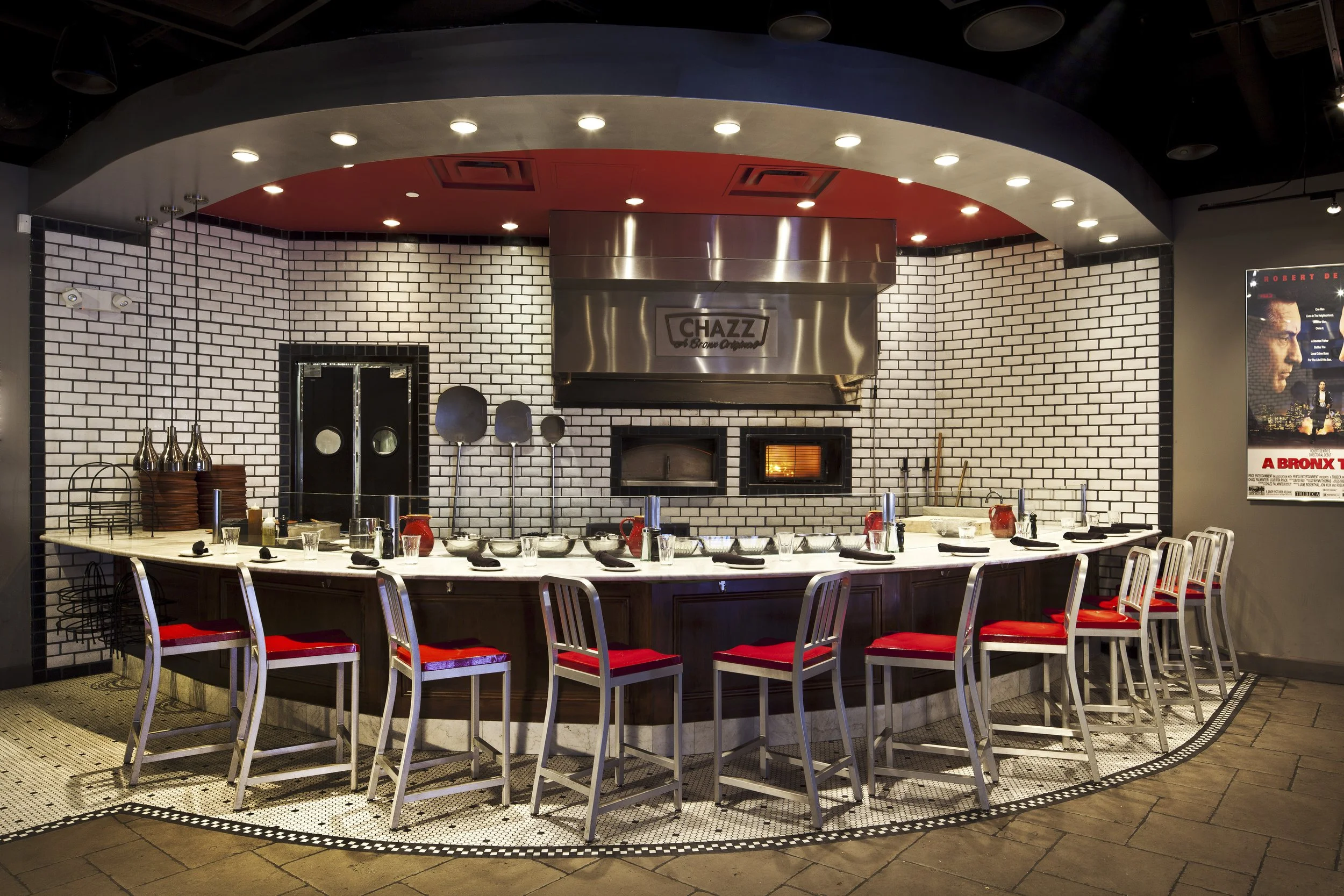 A restaurant with a curved bar counter, white brick wall background, and modern decor. There are chairs with red cushions around the counter, and a pizza oven behind it. The ceiling has recessed lighting, and a movie poster is on the right wall.