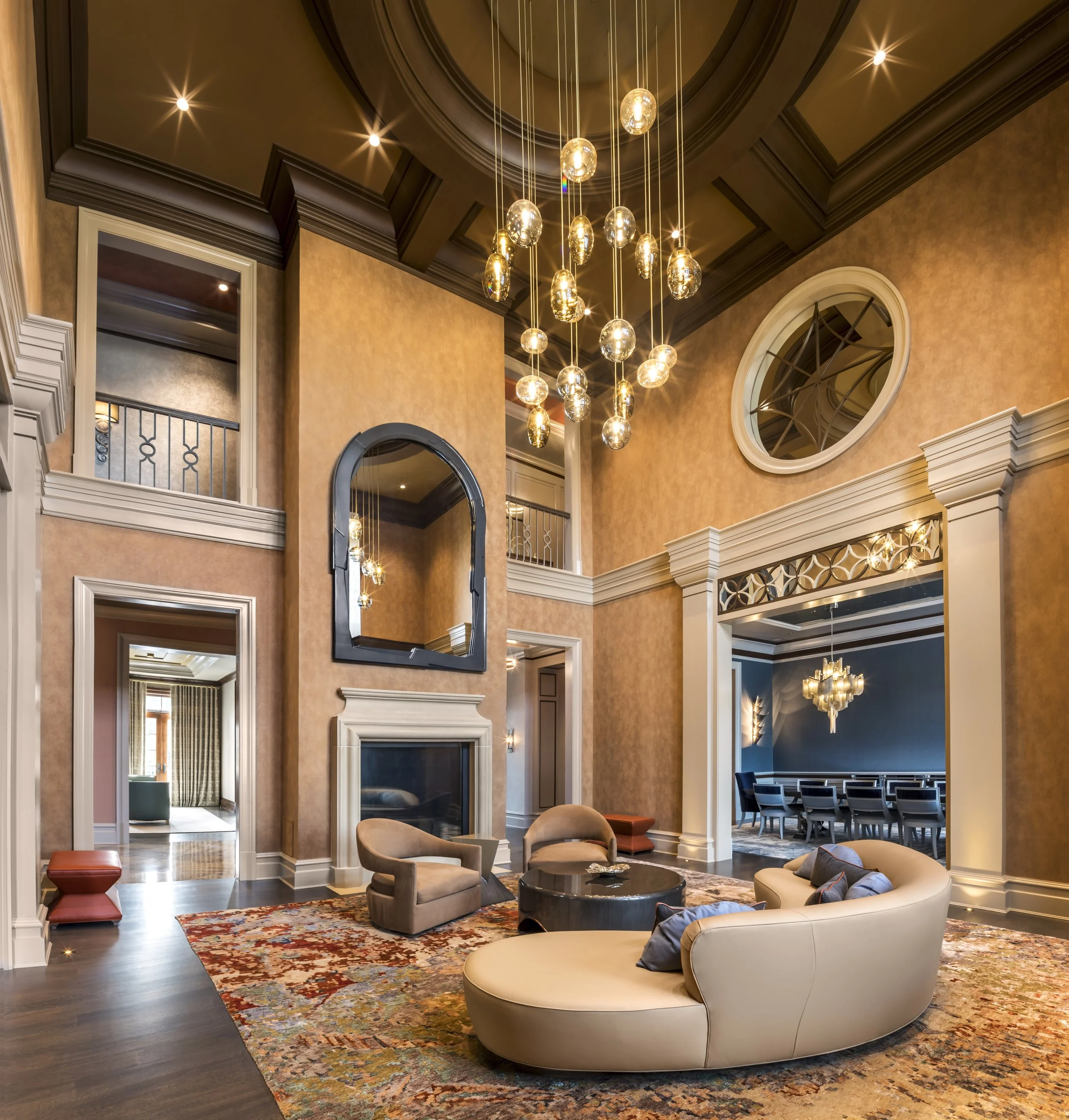 Spacious living room with high ceilings, a modern chandelier, a fireplace, a round beige sofa with cushions, two beige armchairs, a coffee table on a colorful rug, and decorative mirrors on the walls.