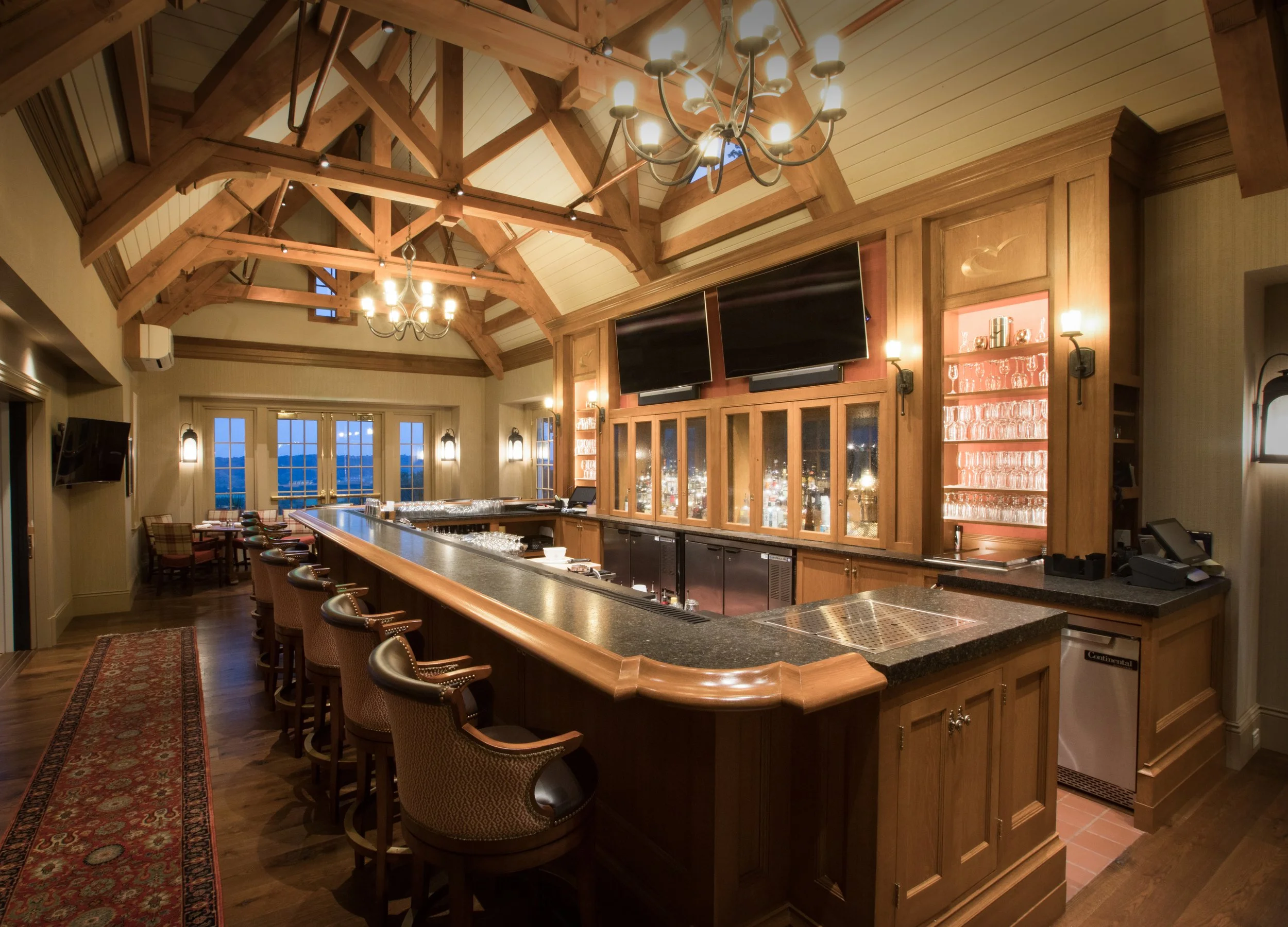 Interior of a cozy bar with a curved wooden counter, barstools, and a high vaulted ceiling with exposed wooden beams. There are two large screens above the bar, with lighting fixtures and glassware on shelves behind the counter.