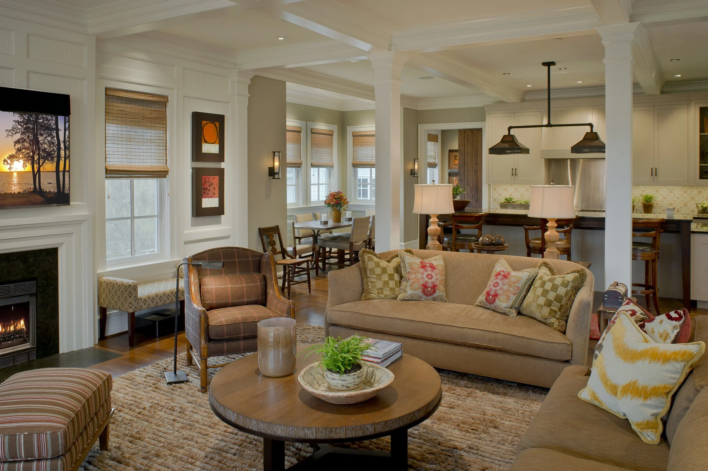 A cozy living room with beige sofas, an armchair, a wooden coffee table with a plant, candles, and books, a fireplace, wall-mounted TV, and large windows with blinds. An open-concept kitchen and dining area are visible in the background.