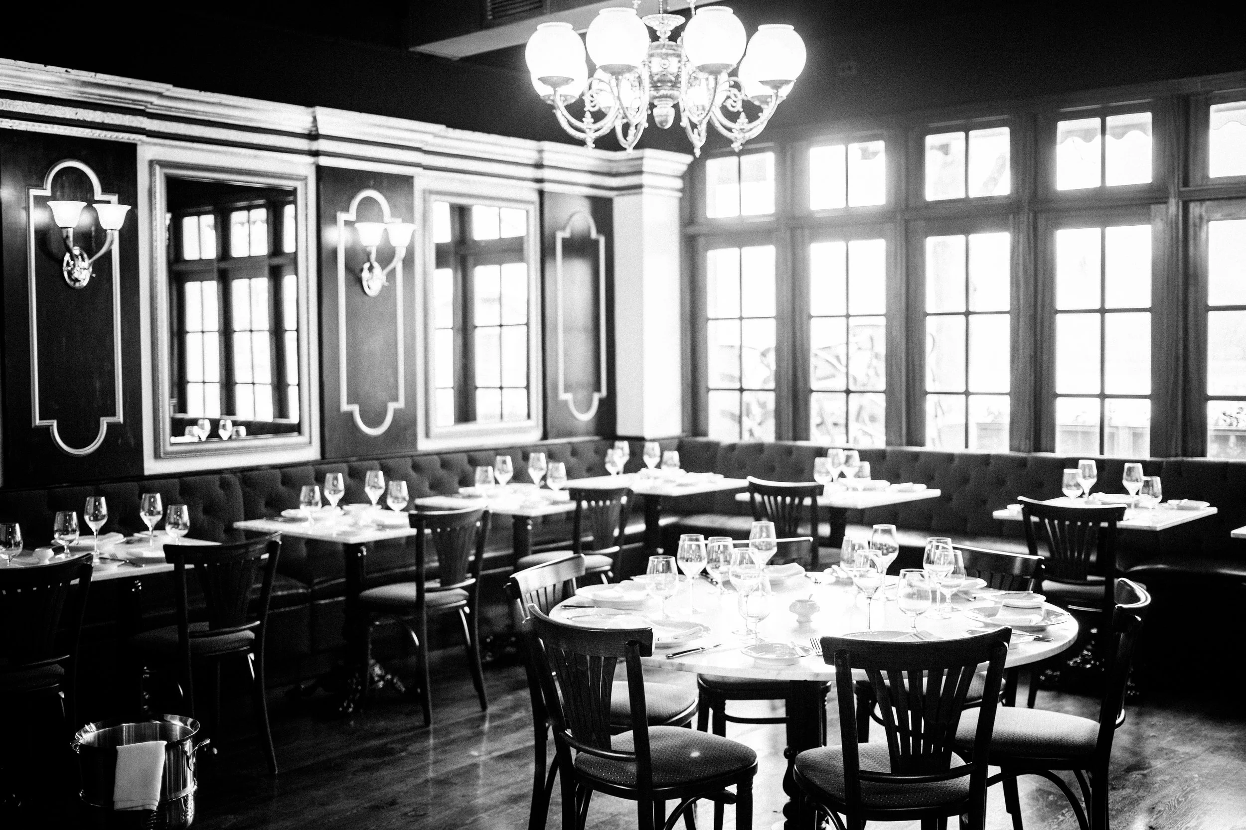 Black and white photo of a restaurant interior with large windows, wooden furniture, a chandelier, and wall sconces.