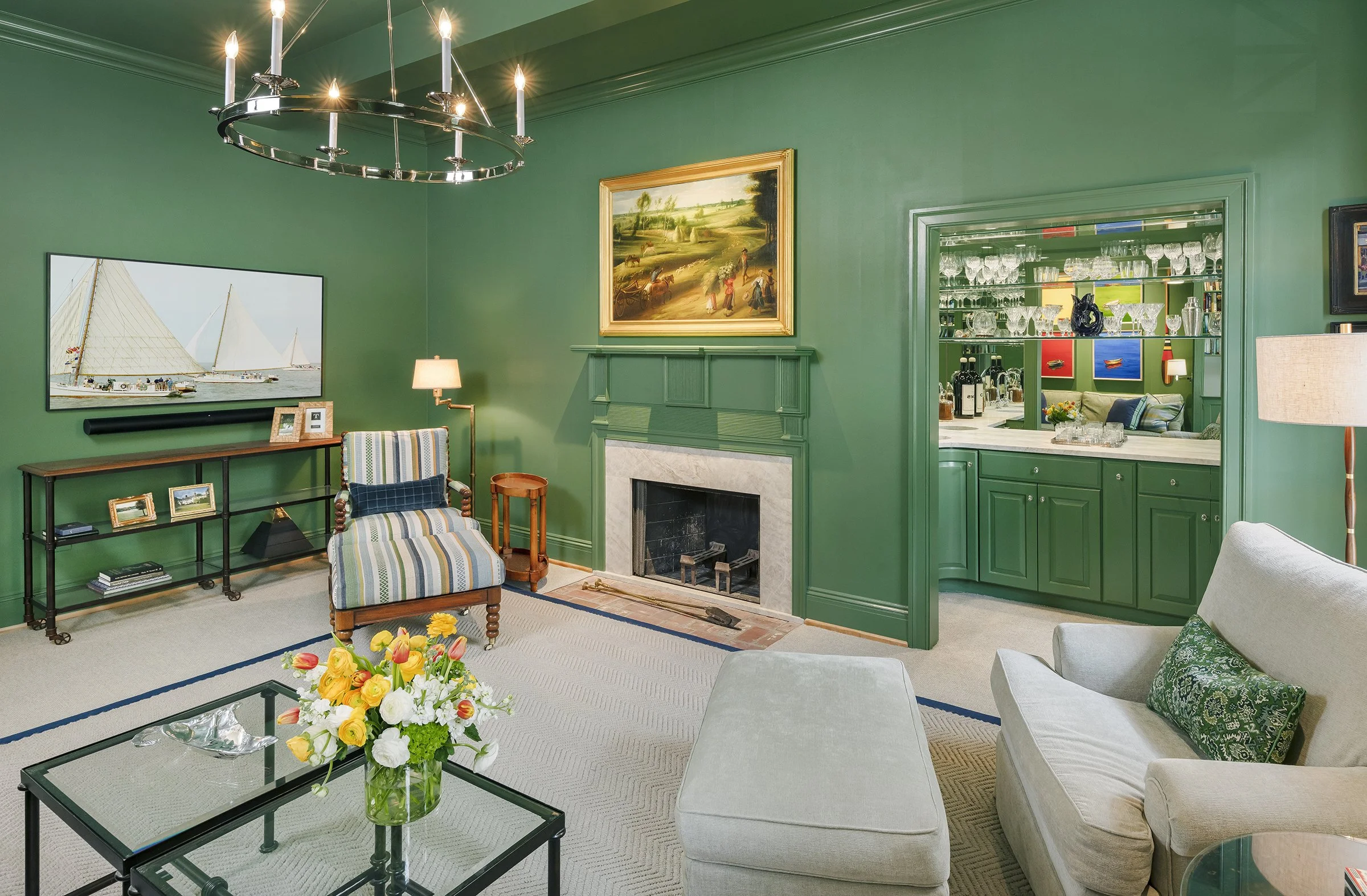 Living room with green walls, a fireplace, a striped armchair, a white sofa, a glass coffee table with a flower arrangement, and a media console with framed photos. There is artwork above the fireplace and a chandelier hanging from the ceiling. An op