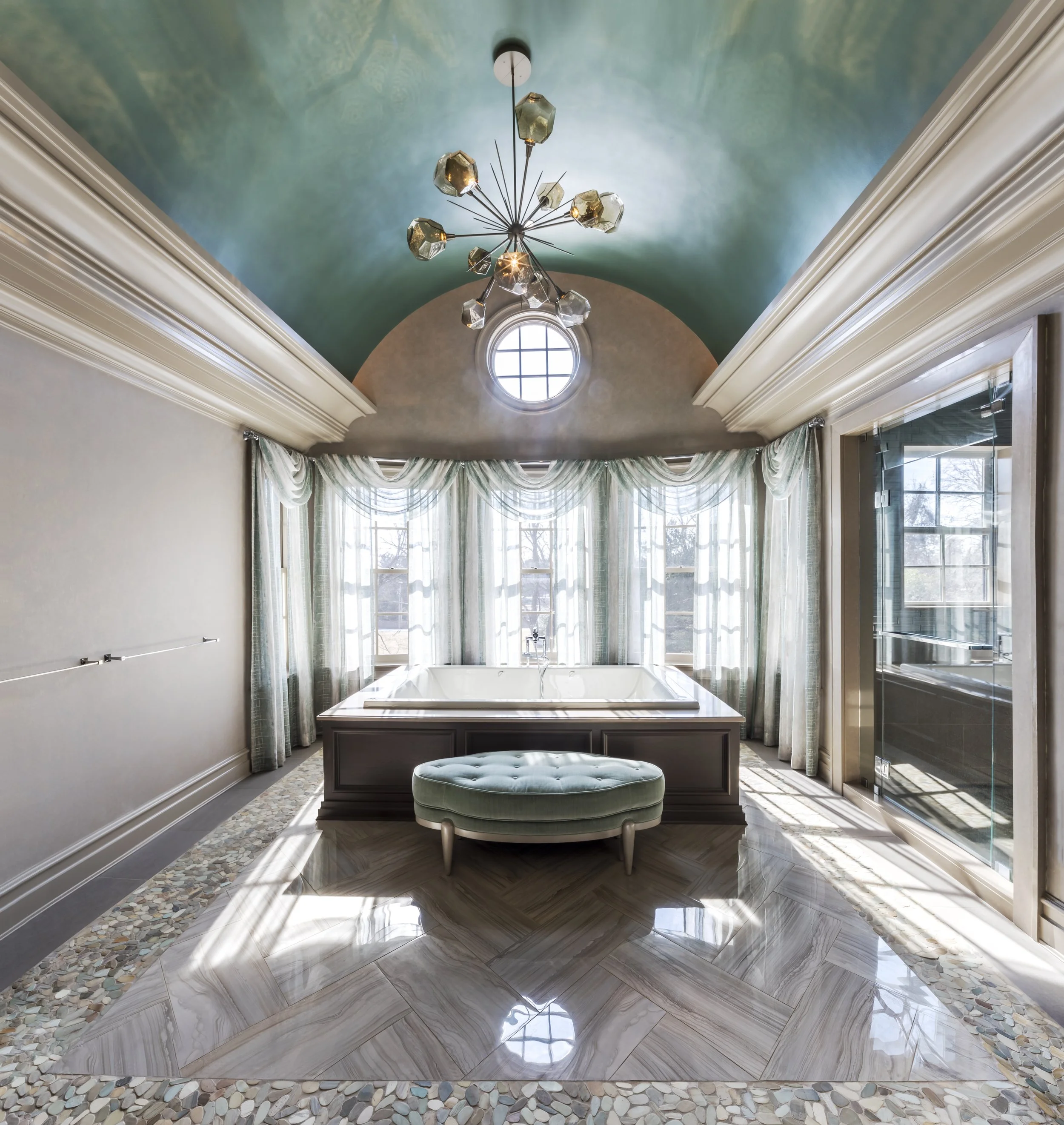 Luxurious bathroom with a bathtub beneath large windows, a round window in the ceiling, an elegant chandelier, and a padded stool.