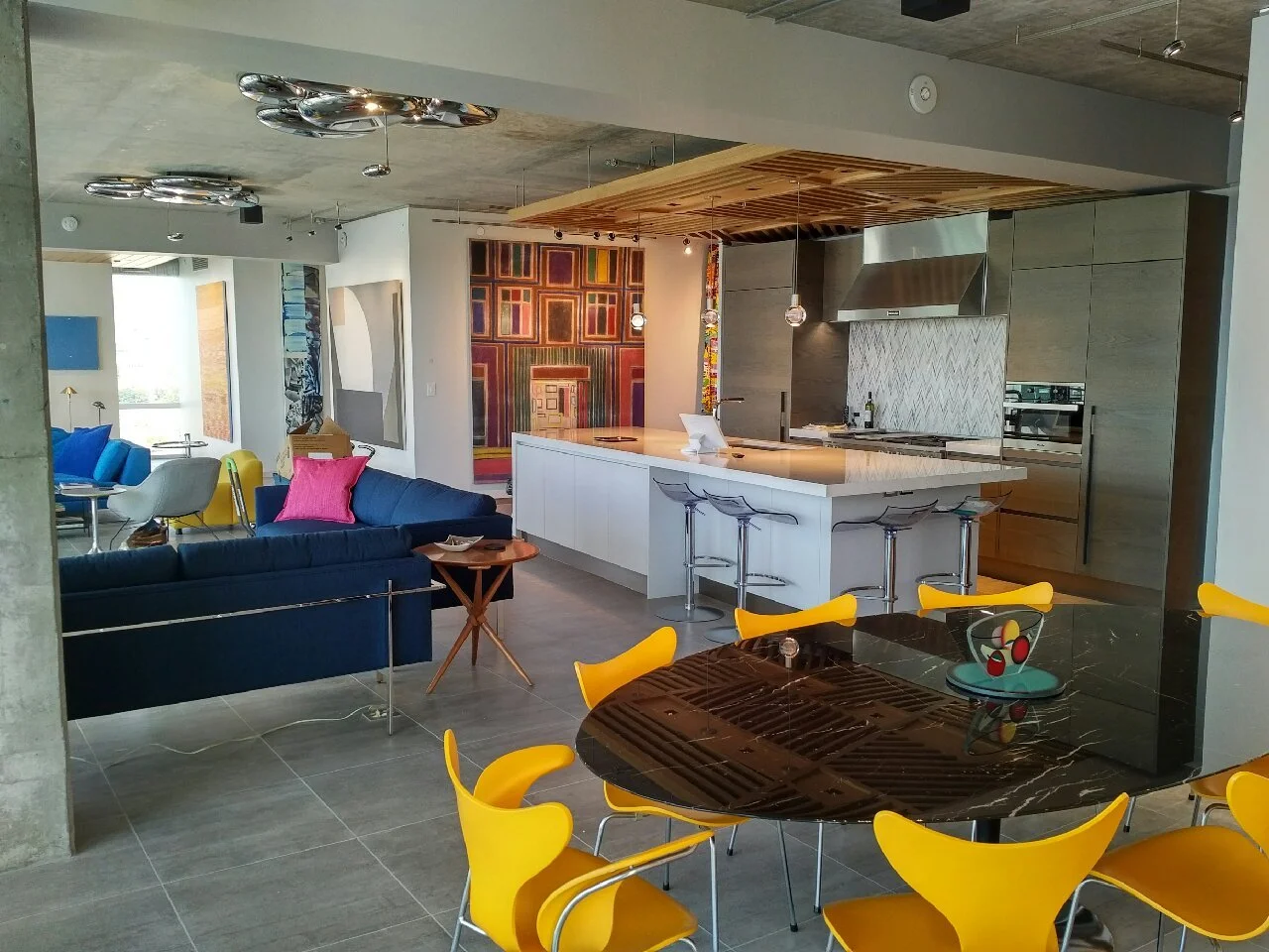 Open-concept modern living space with a kitchen island, colorful chairs around a black marble table, a navy blue sofa with pink pillows, and abstract artwork on the walls.