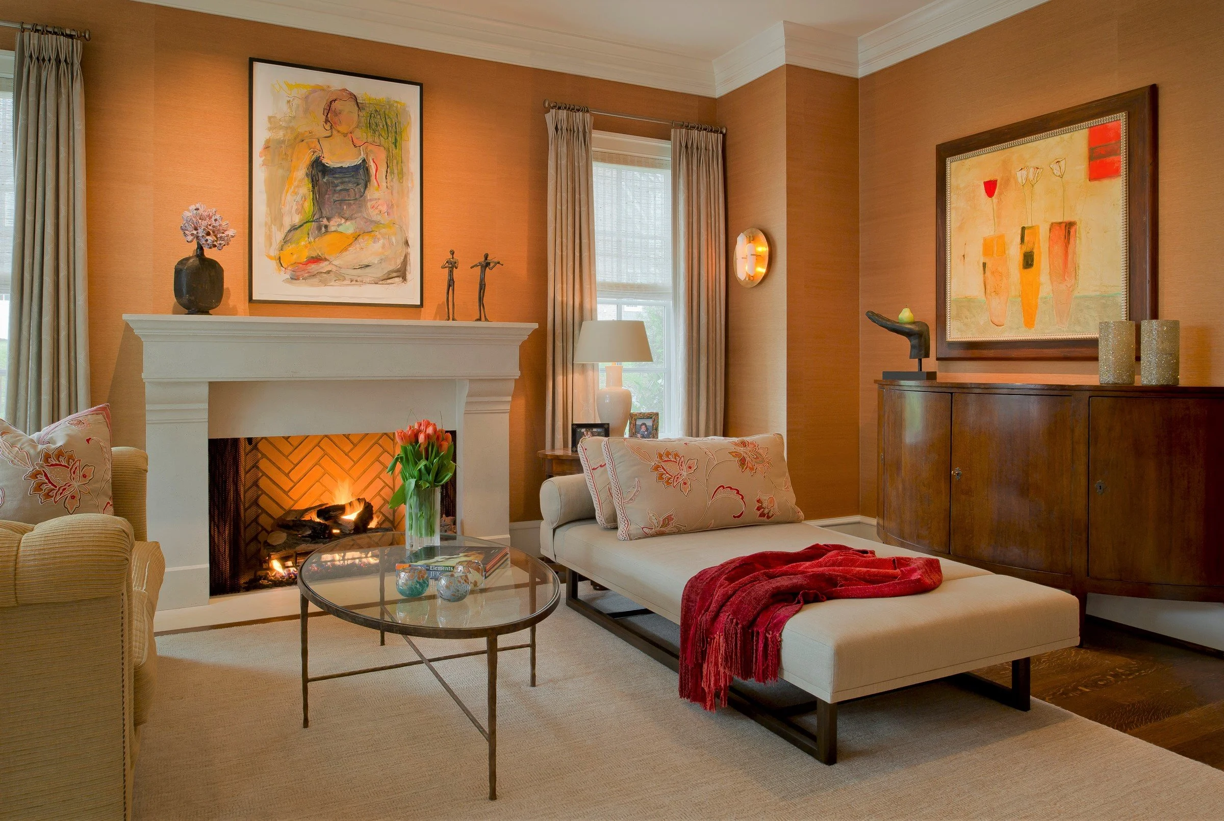 A living room with a fireplace, a glass coffee table with flowers, a white chaise lounge with pillows and a red throw, a side table with a lamp, artwork, and decorative items.