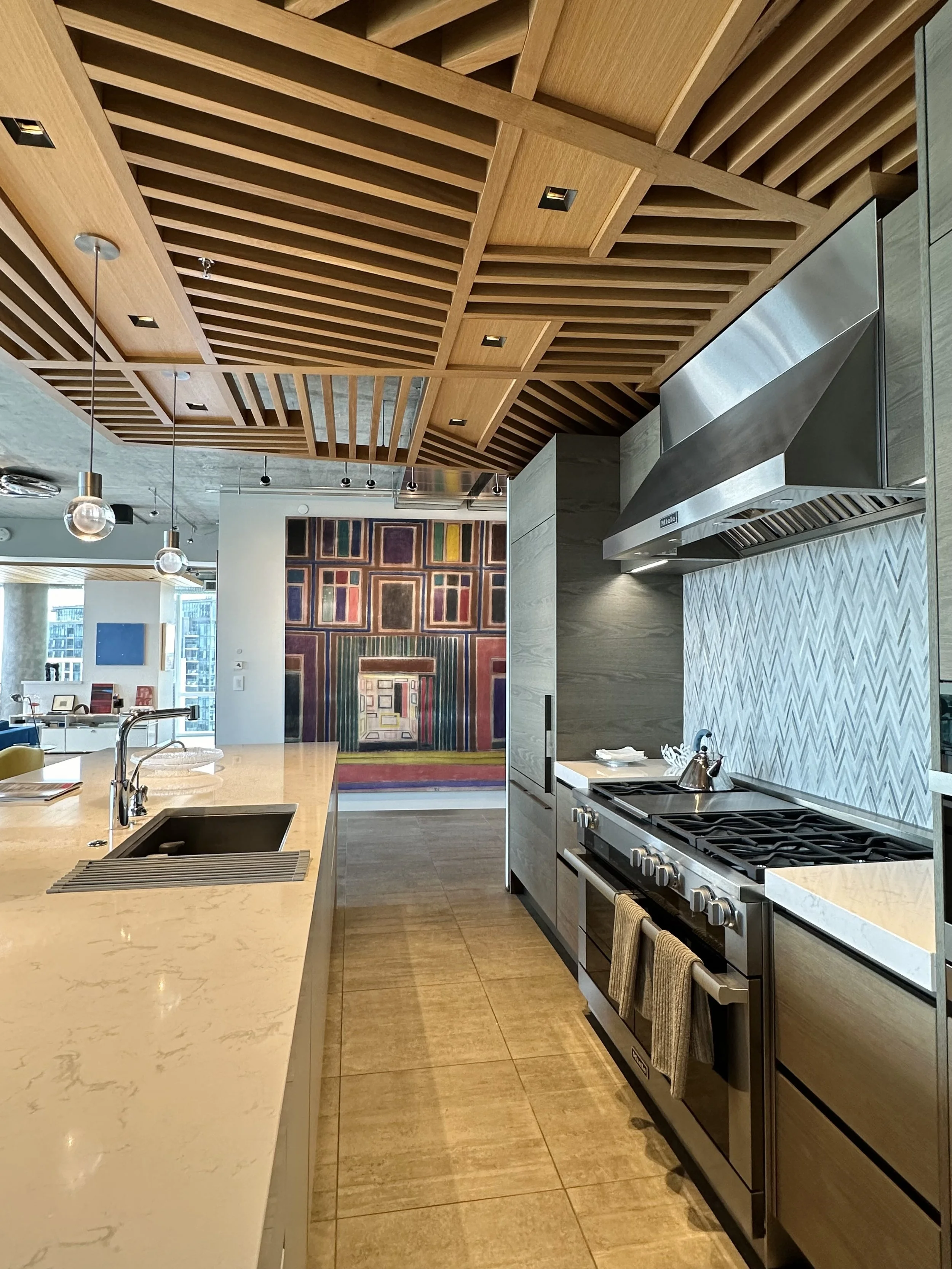 Modern kitchen with marble countertops, stainless steel appliances, wooden ceiling design, and abstract colorful artwork on the wall.