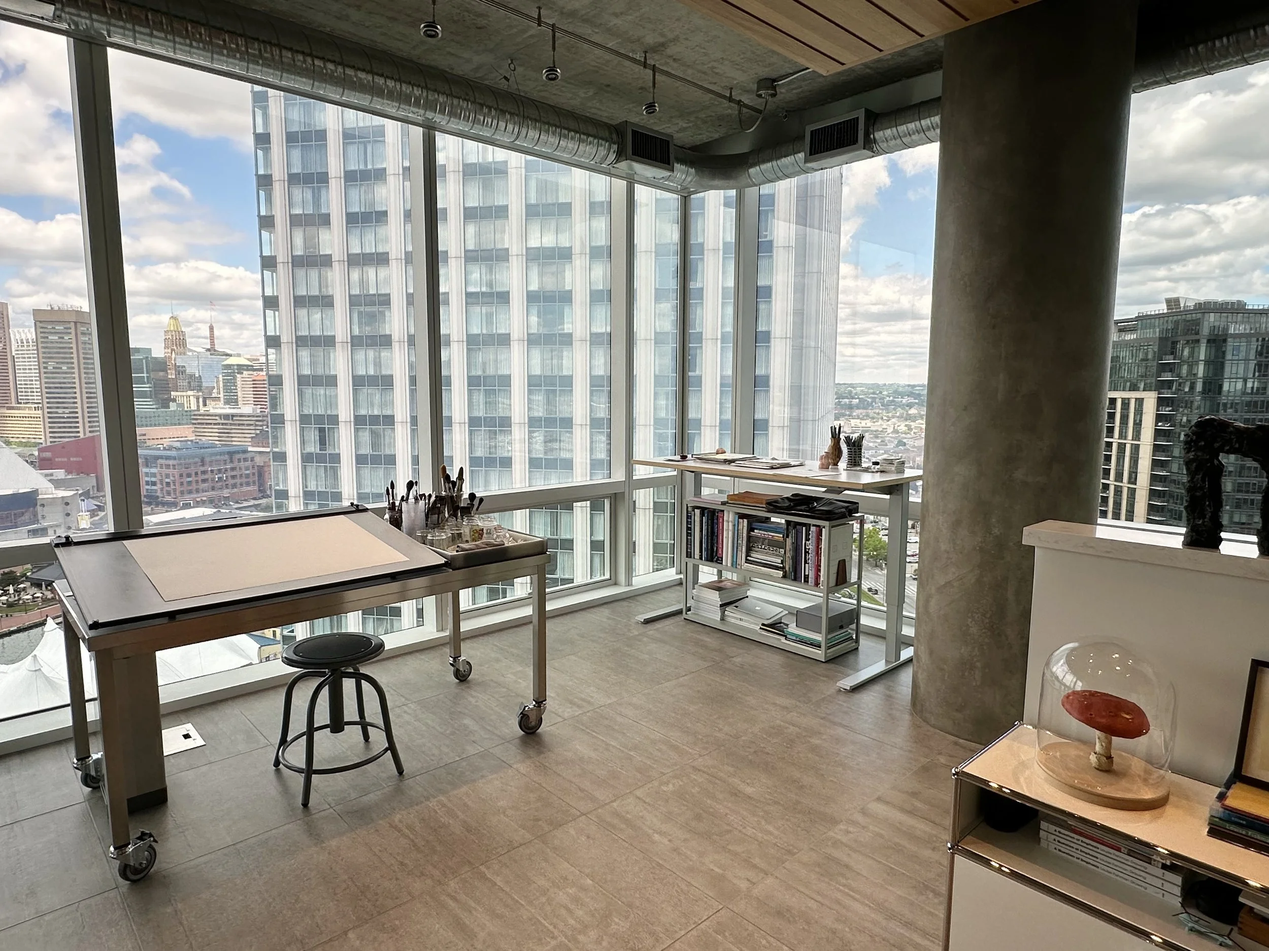 Modern office space in a high-rise building with large windows overlooking a city skyline, featuring a drafting table with art supplies, bookshelves, and a display case with a mushroom sculpture.