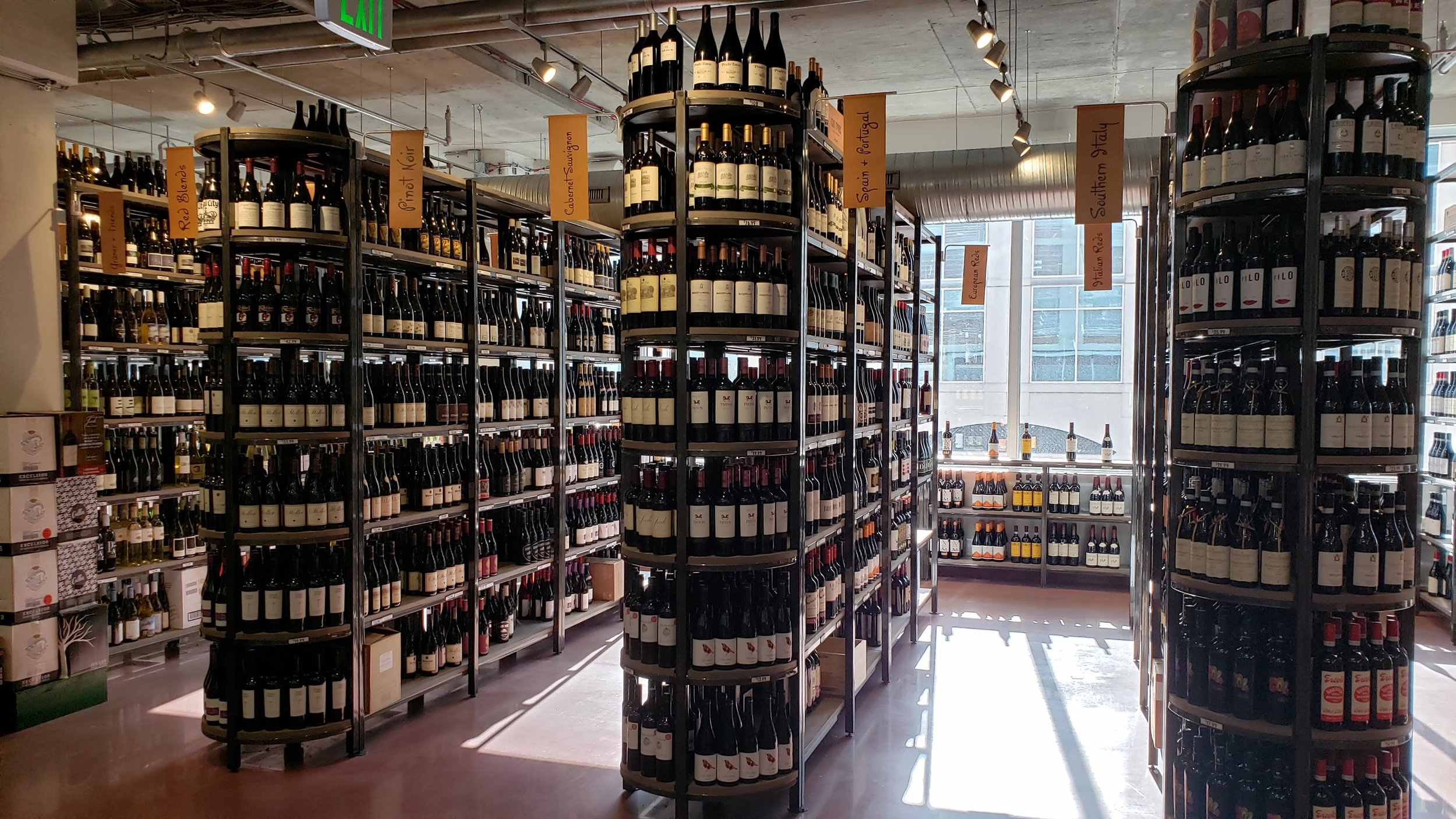 Interior of a wine store with shelves filled with bottles of wine, illuminated by natural light from large windows at the back.