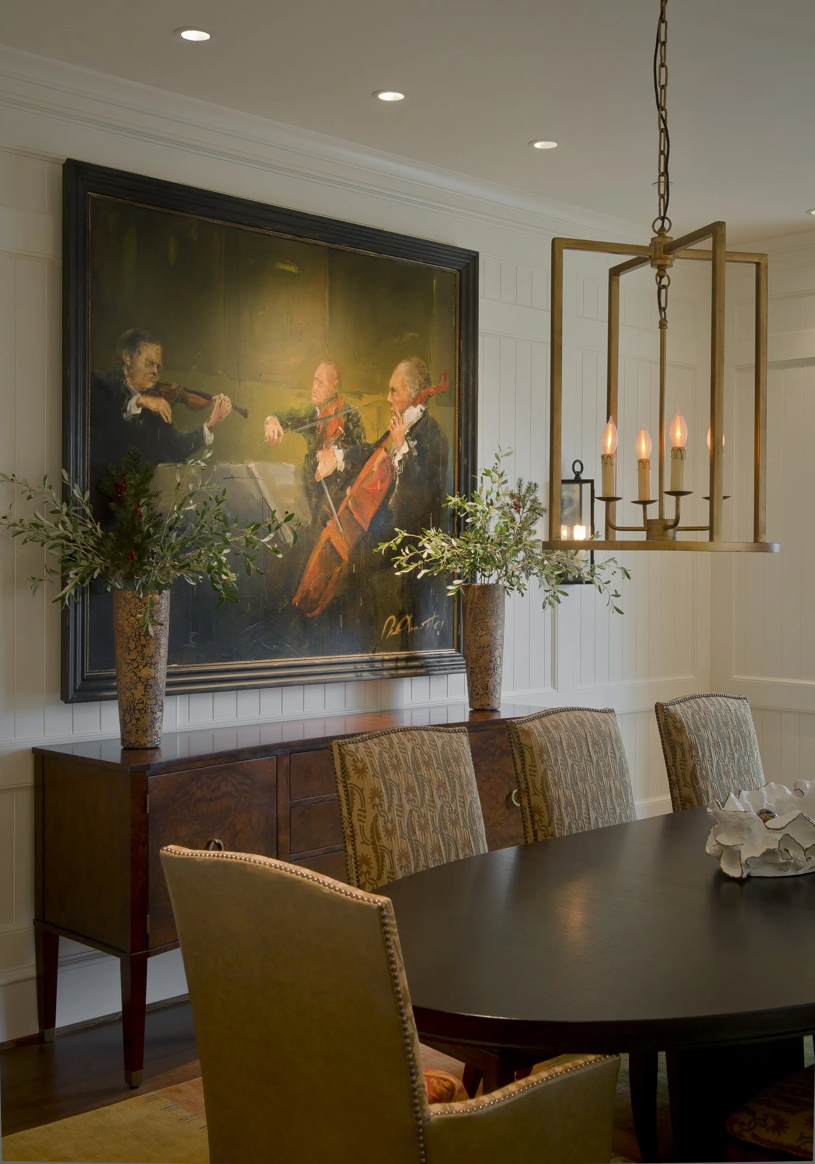 An elegant dining room with a large painting of three musicians playing string instruments, two tall floral arrangements in decorative vases on a sideboard, and a modern chandelier with candle-shaped lights hanging above a dark wooden dining table su
