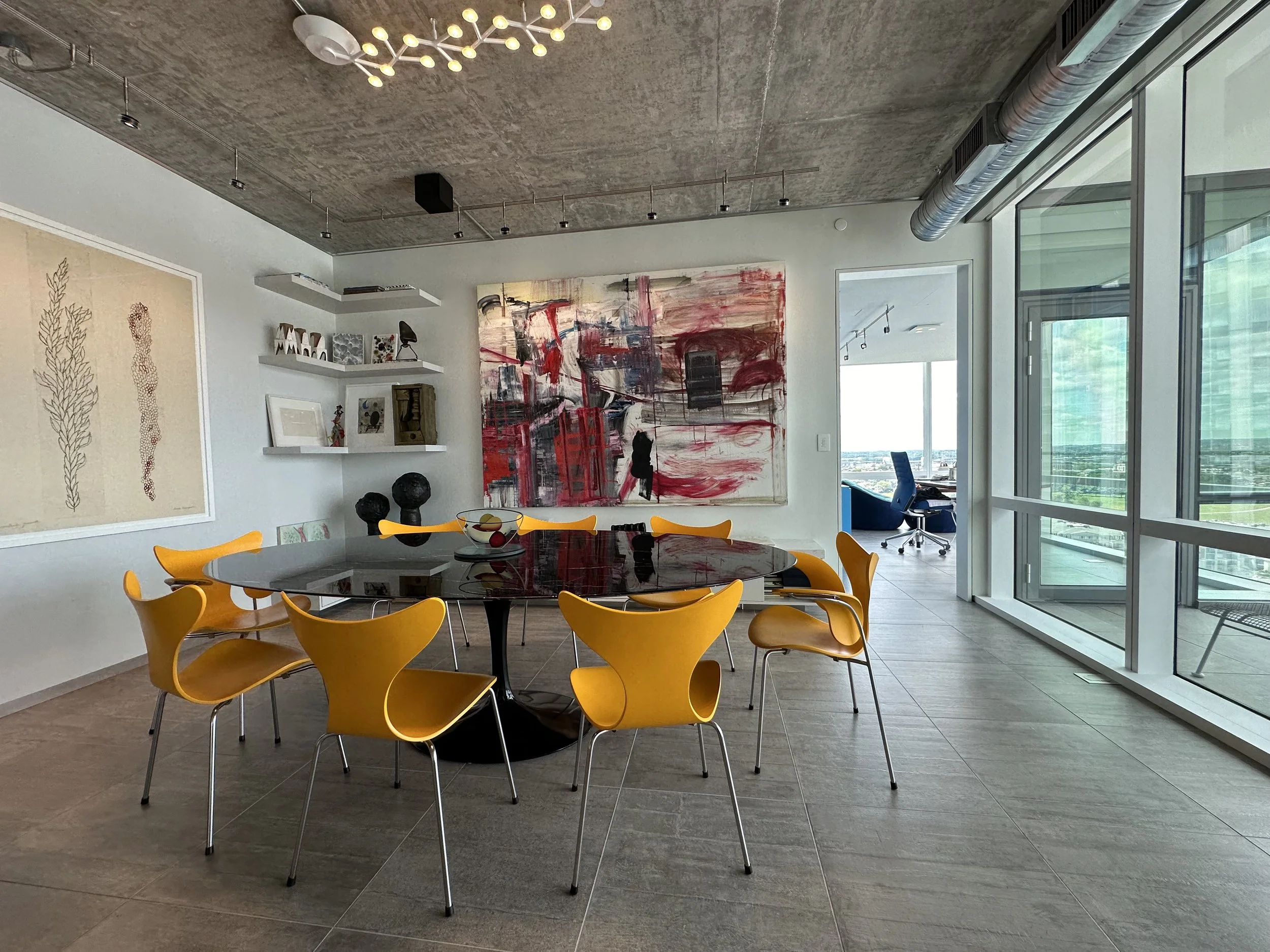 Modern dining area with a black round table and yellow chairs, abstract paintings on the walls, large windows with a view, and a concrete ceiling.