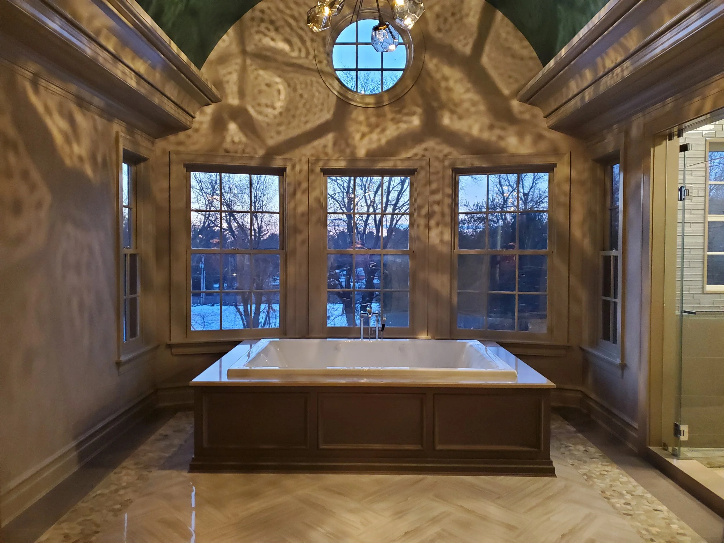 A luxurious bathroom with a built-in bathtub beneath large windows, wooden paneling, and a decorative ceiling with a circular window and hanging light fixture.
