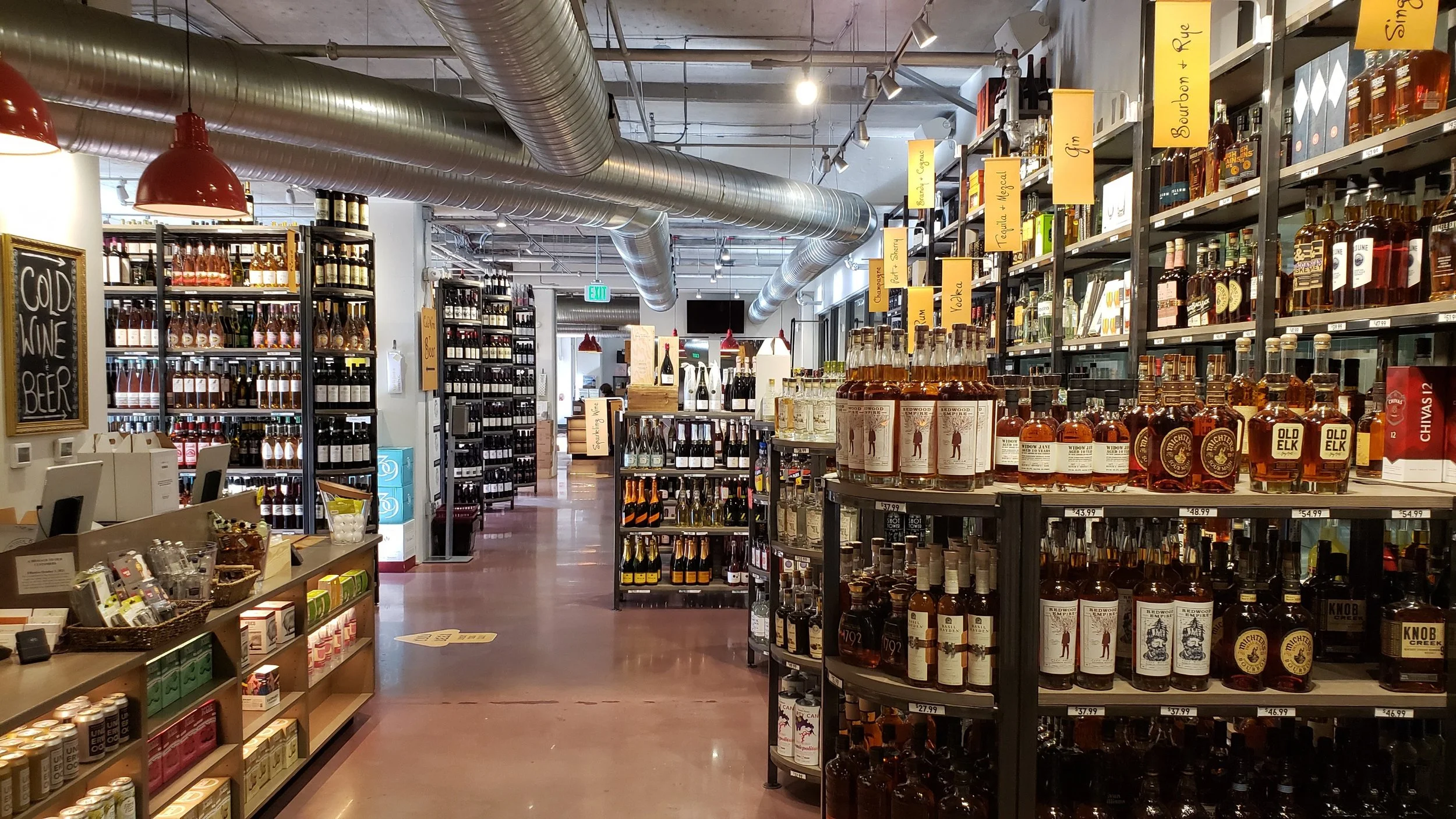 Interior of a liquor store with shelves stocked with bottles of alcohol, signs indicating different liquor categories, and a counter area at the front.
