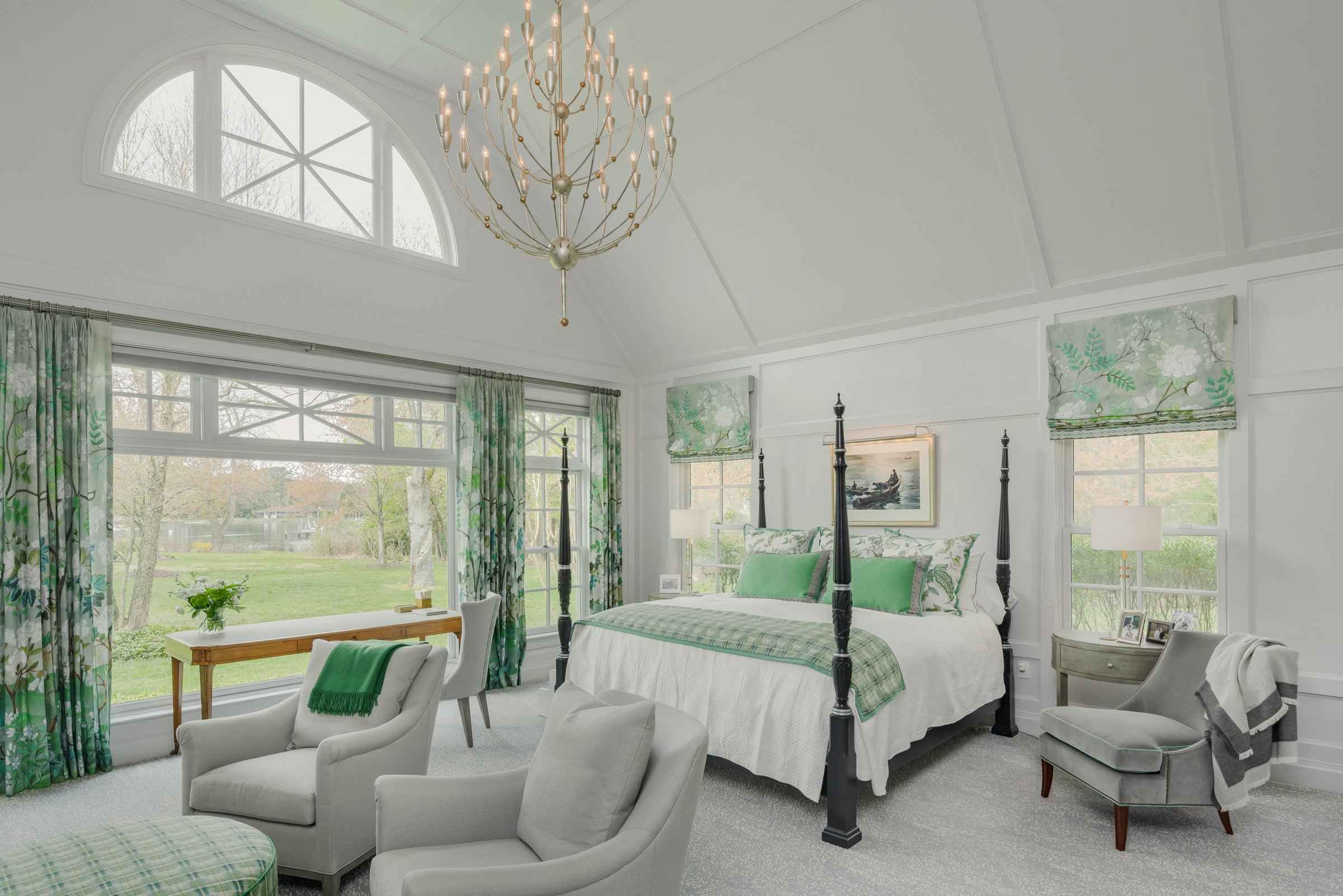 Bright bedroom with a high vaulted ceiling, large arched window, and floral curtains. Contains a four-poster bed with green and white bedding, a chandelier, a sitting area with armchairs, and a lake view outside.