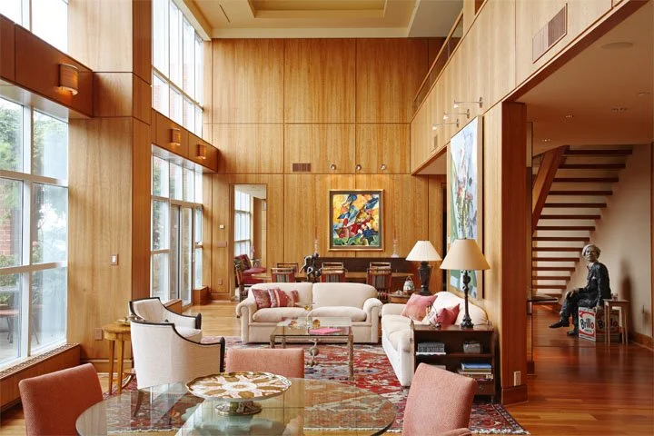 Spacious living room with wooden walls and large windows, furnished with sofas, armchairs, coffee tables, and colorful pillows, decorated with artwork and lamps.