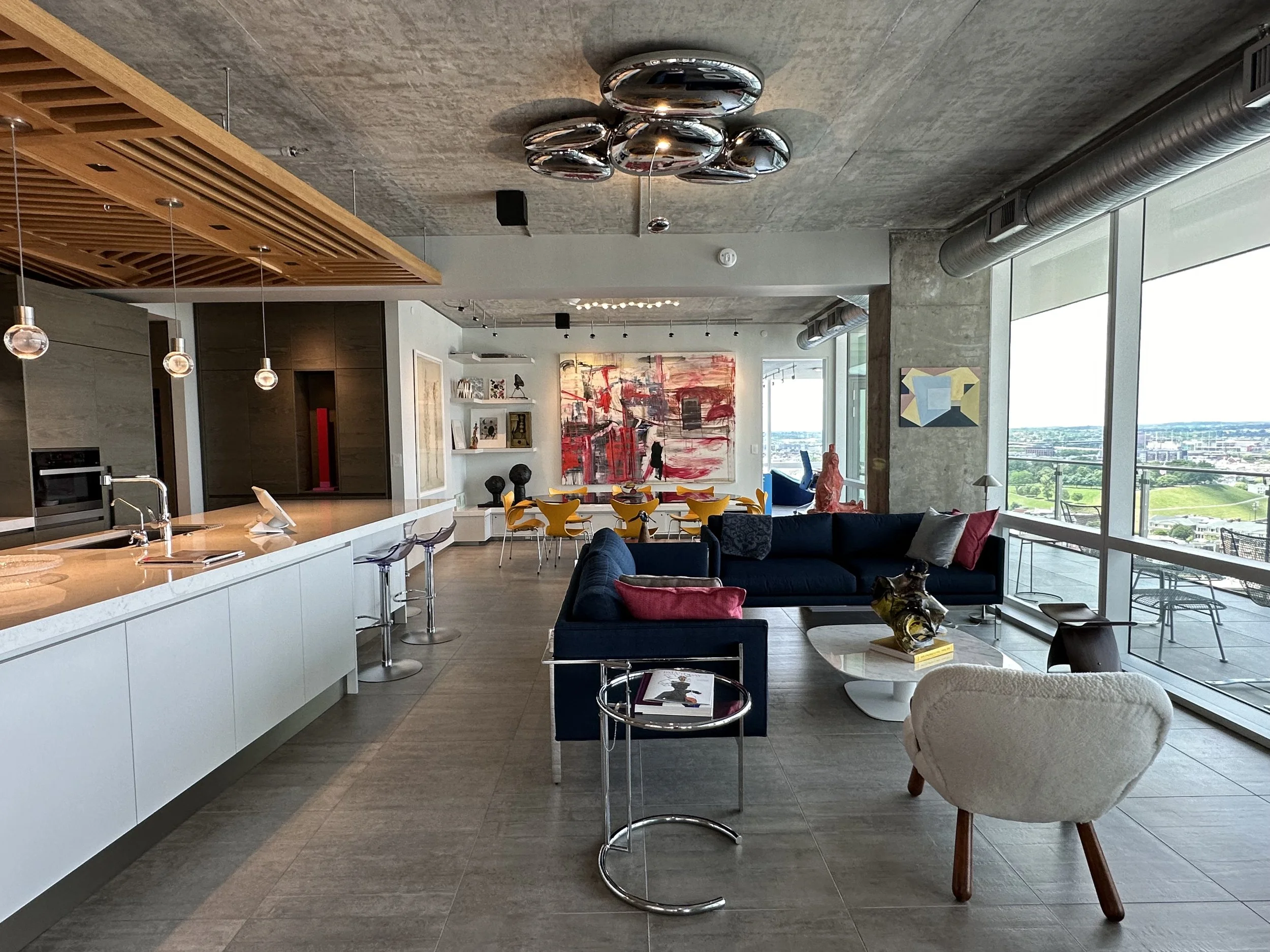 Modern open-concept living space with a kitchen, dining area, and lounge. The space features large windows, contemporary furniture, abstract art, and a view of the city outside.