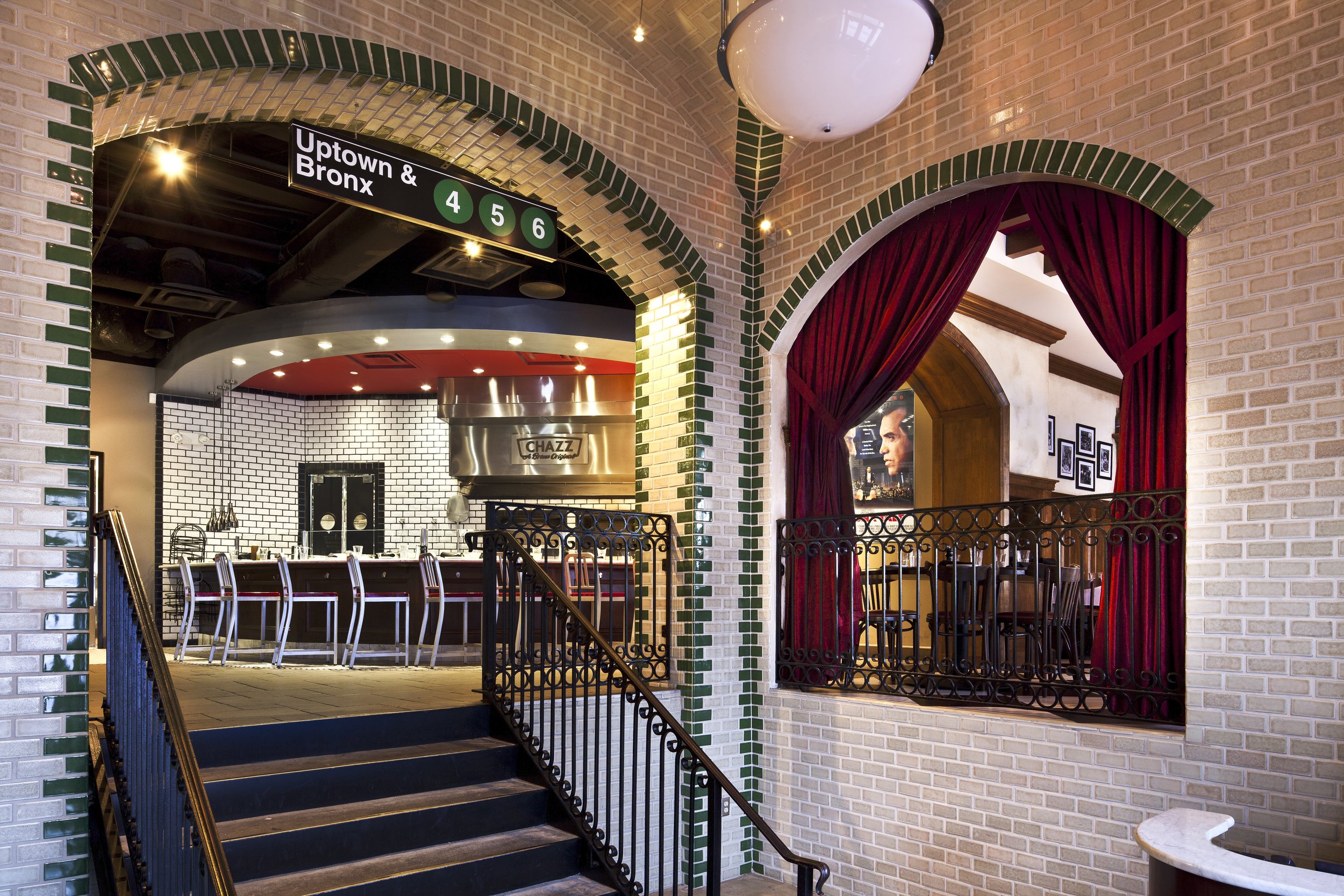 Interior of a restaurant or bar, seen from the staircase, with a sign overhead indicating subway lines 4, 5, and 6 for uptown and Bronx. There is a bar area with white chairs and a black and white tile kitchen area behind it, as well as a separate di