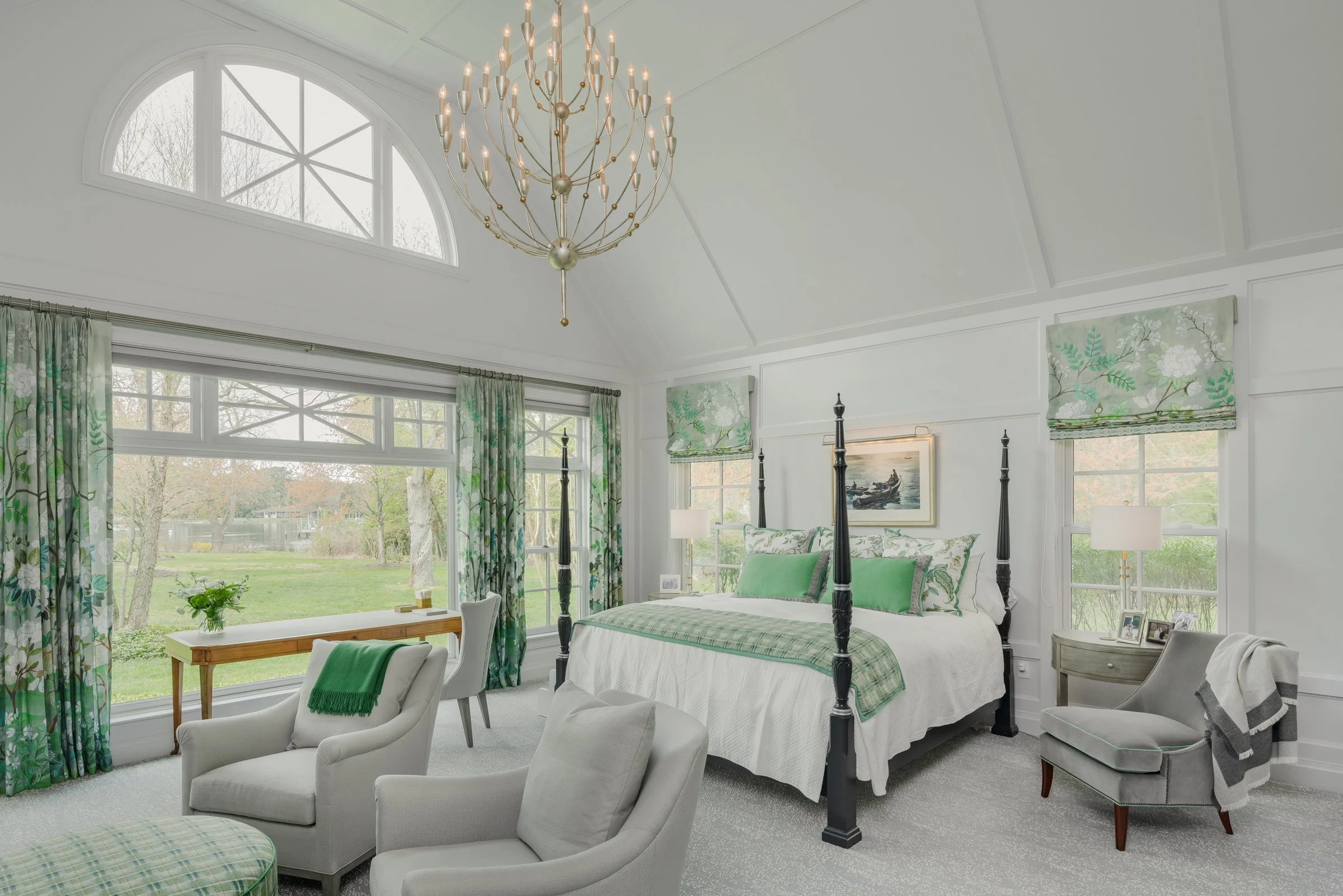 Bright bedroom with a high ceiling, large arched window, chandelier, four-poster bed with green accents, white walls, and green curtains with floral patterns.