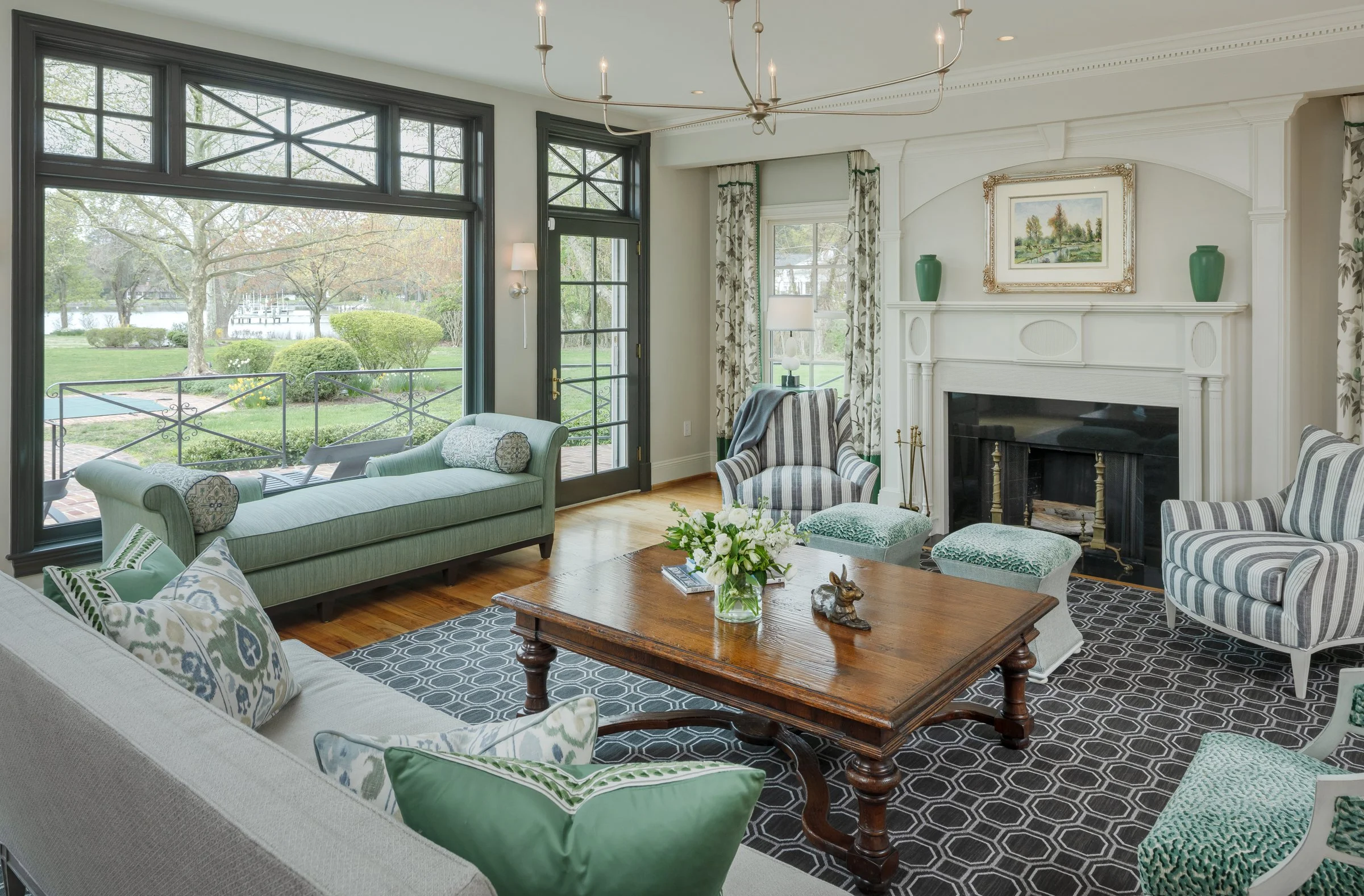 Living room with large windows overlooking a garden, white fireplace, striped armchairs, a wooden coffee table with a flower arrangement, and patterned cushions.