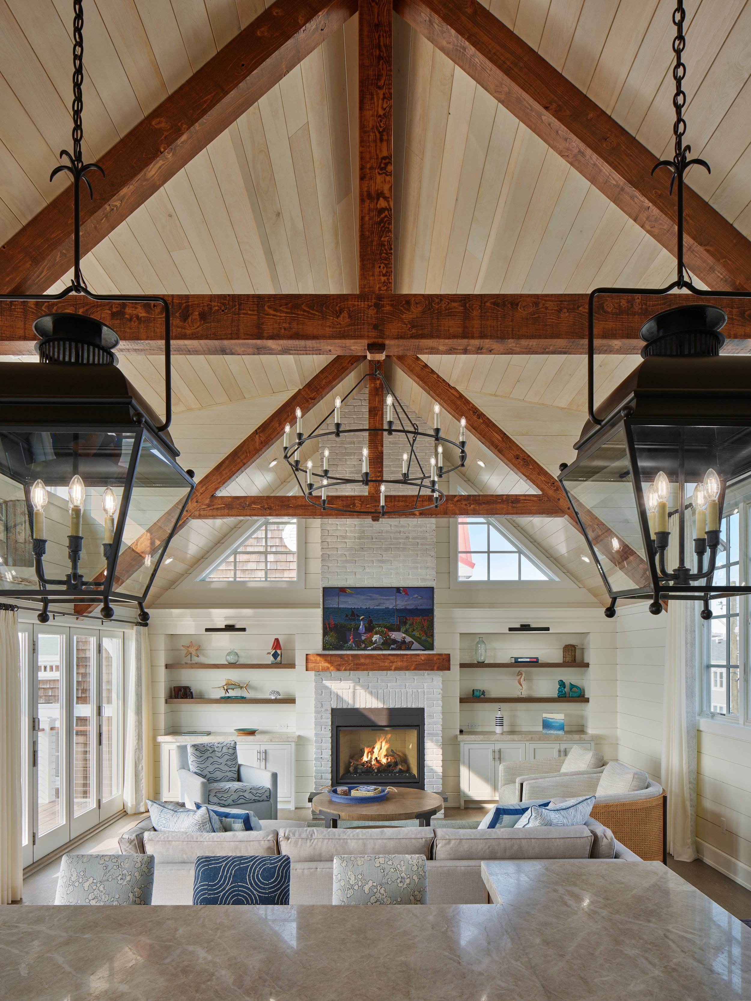 Living room with white brick fireplace, wall-mounted TV, and vaulted ceiling with wooden beams. Seating includes a sofa, armchairs, and decorative pillows. Surrounding shelves hold art and decorations. Large windows and sliding glass door provide nat
