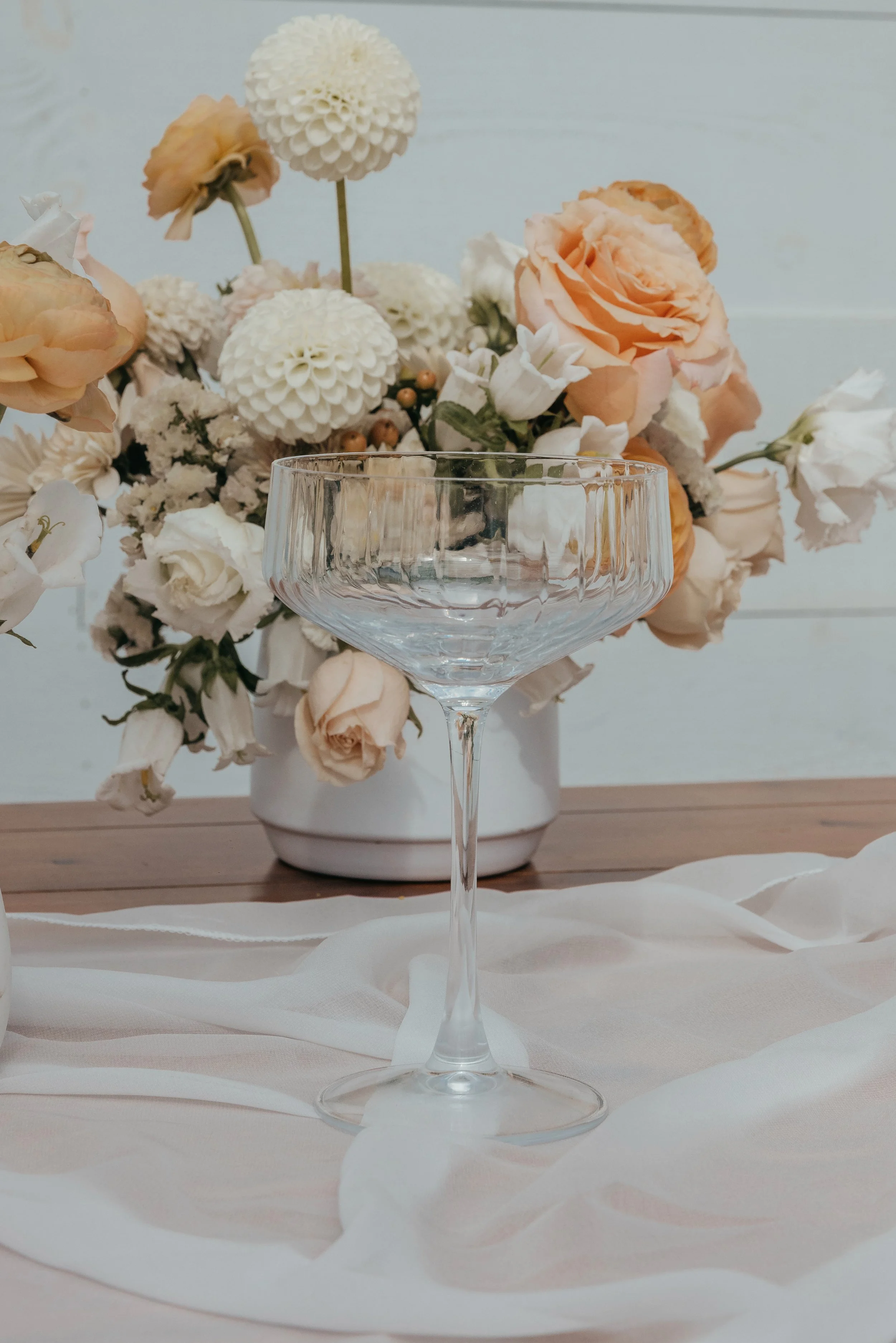 Clear empty cocktail glass in front of a bouquet of peach and white flowers on a wooden table