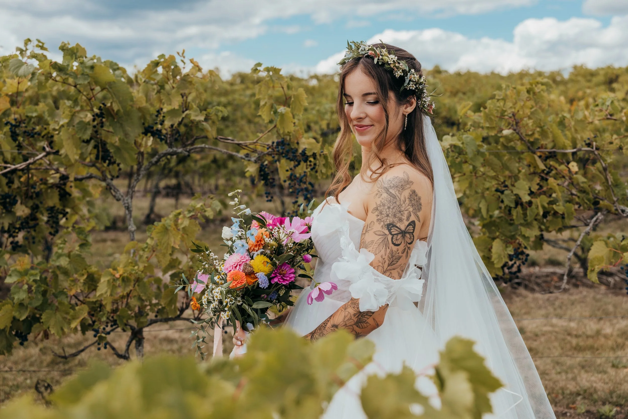 Bridal portrait of a woman holding a colorful bouquet, standing in a vineyard surrounded by grapevines, with a floral crown, lace wedding dress, and veil.