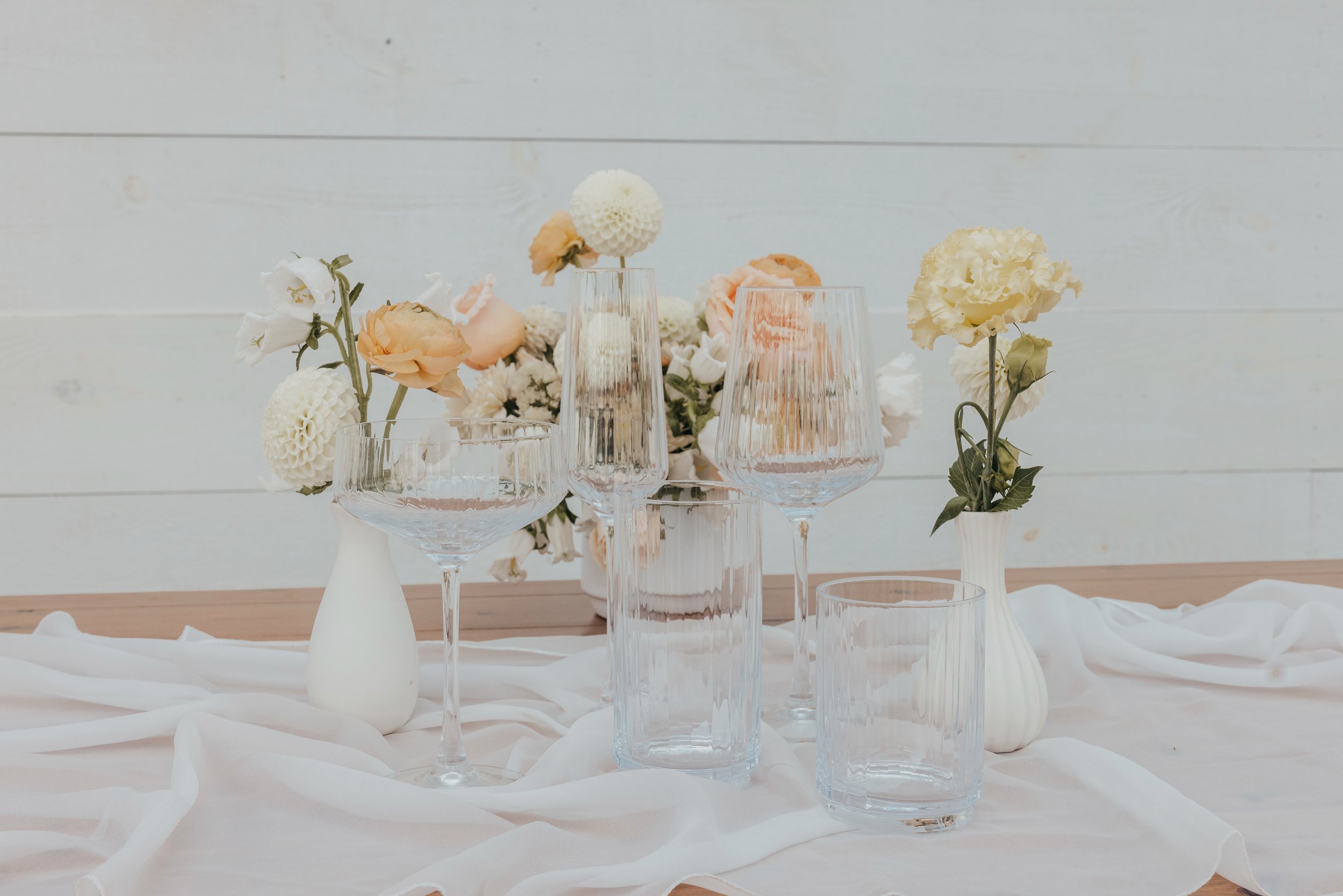 Table setting with glassware and floral arrangements in white vases on a soft fabric, against a light wooden wall background.