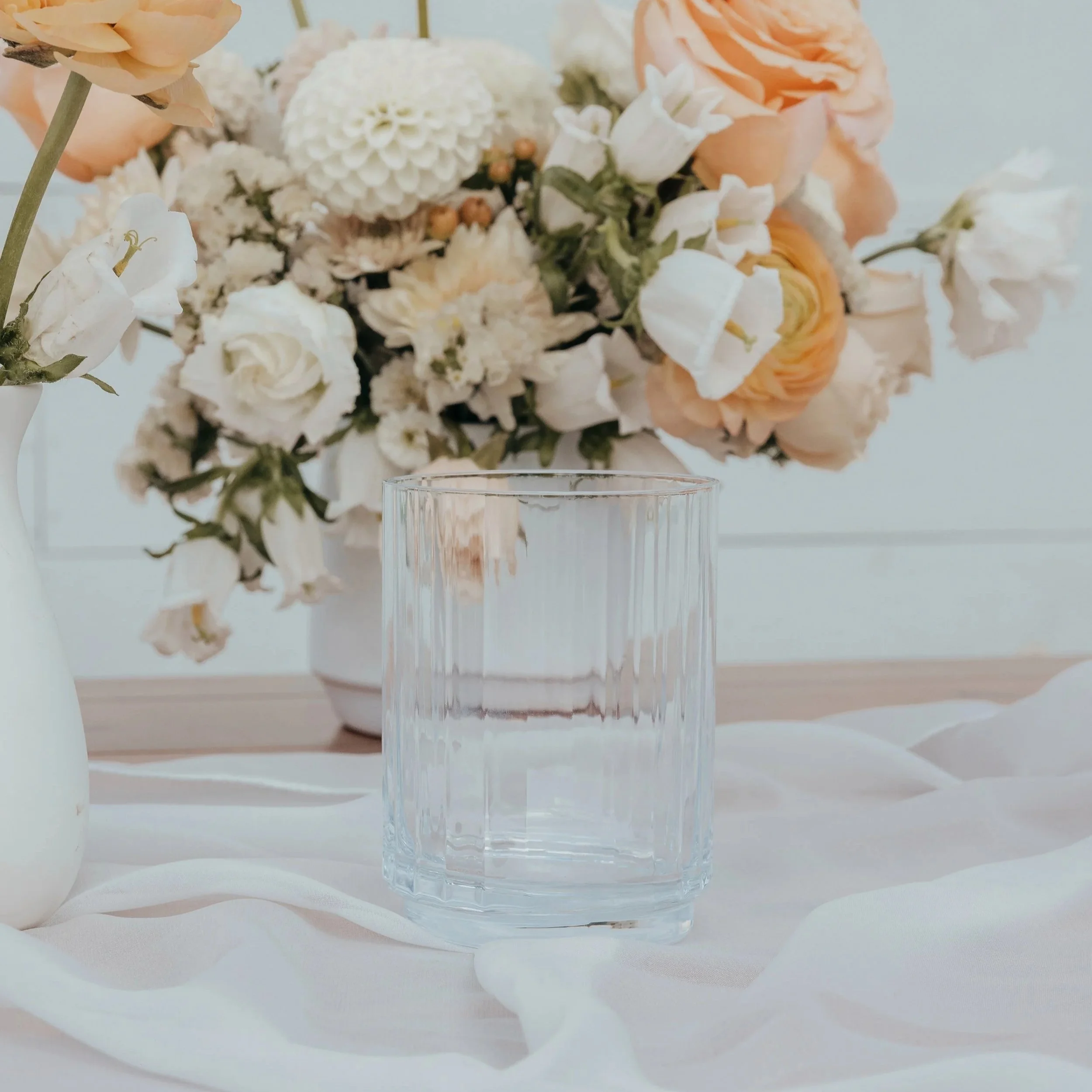 Clear glass of water on a white tablecloth with a floral arrangement of white and peach flowers in a white vase in the background.