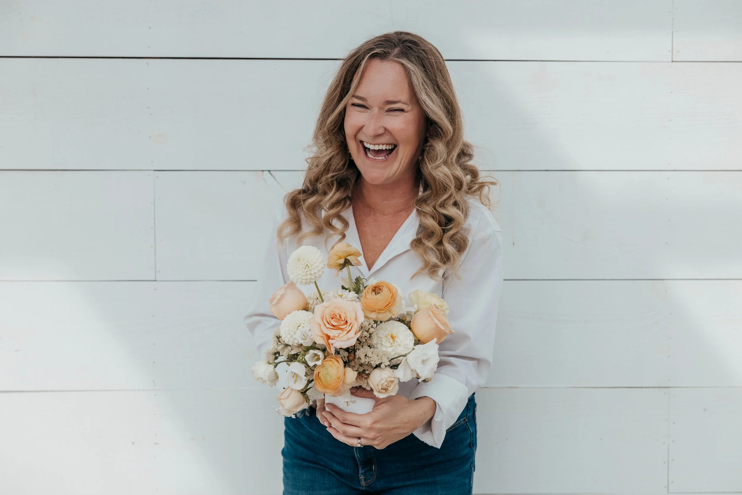 Owner of The Bloom of Lynn, Brittany with curly blonde hair, smiling and holding a bouquet of light-colored flowers in front of a white wooden wall is posing for a head shot.