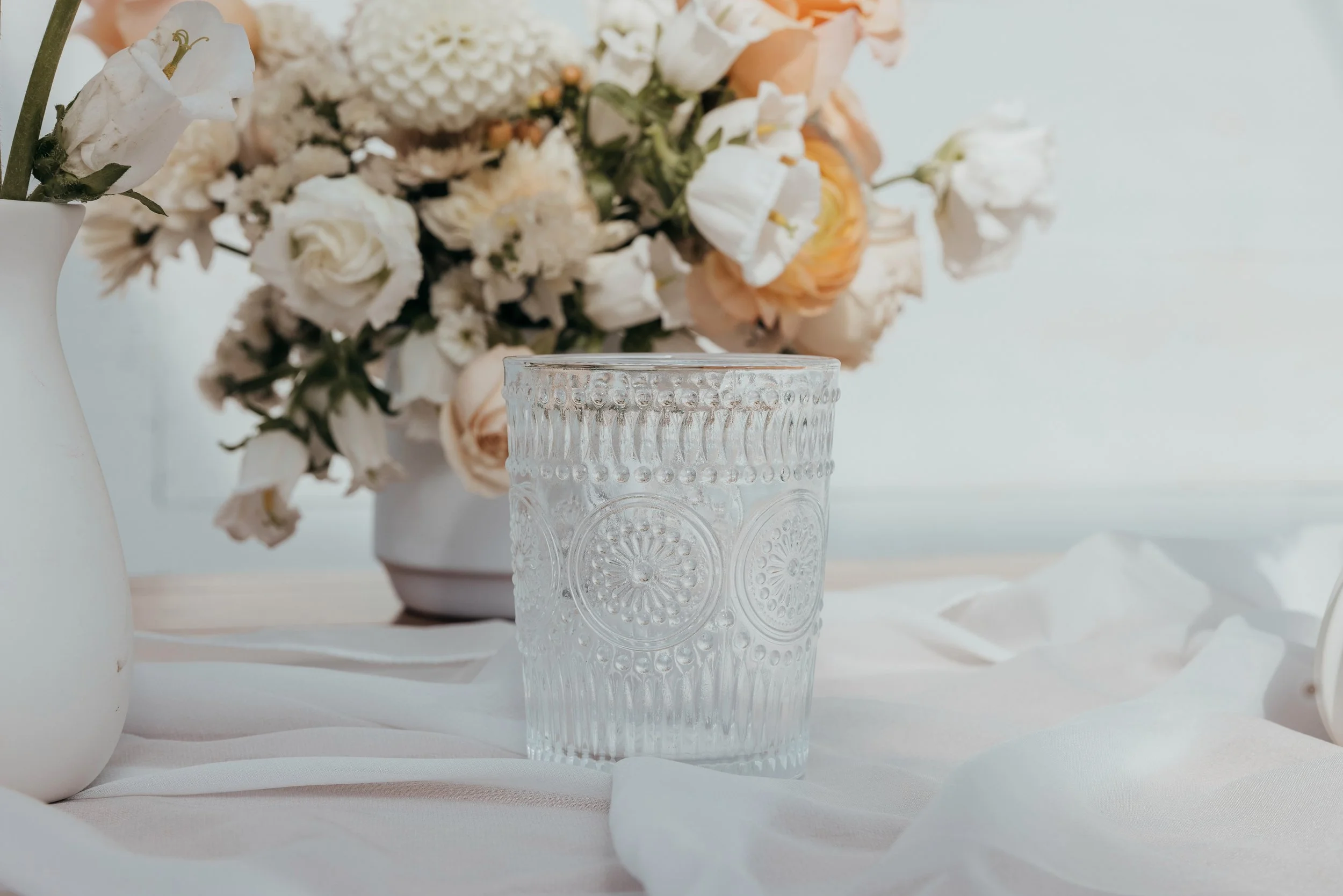 A clear glass with intricate circular patterns on a white surface, surrounded by flower vases with white and peach-colored flowers, with a soft light background.